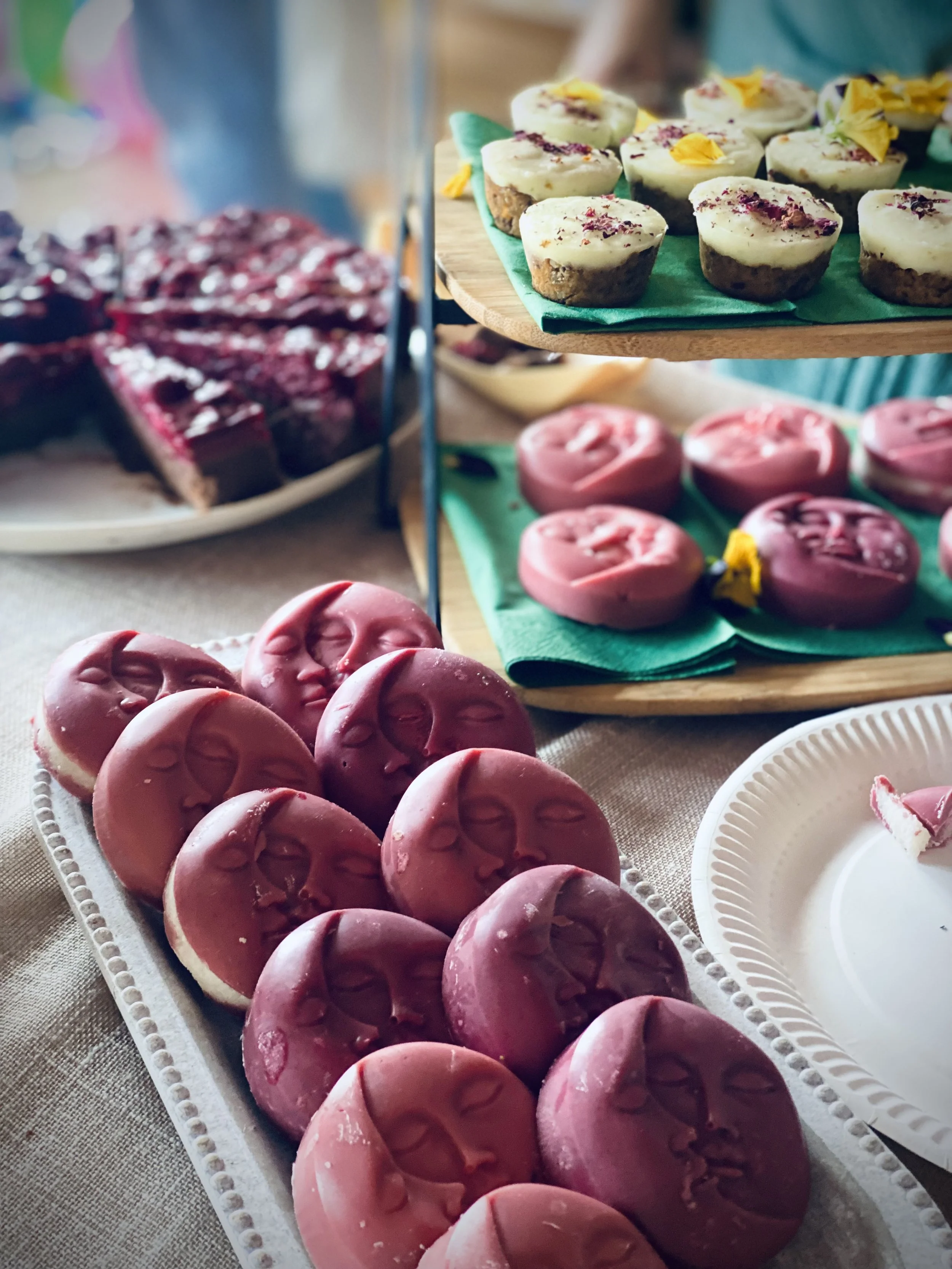 Assorted desserts including pink cookies with face impressions, cupcakes with yellow flowers, and slices of red velvet cake on a table at a dessert buffet.