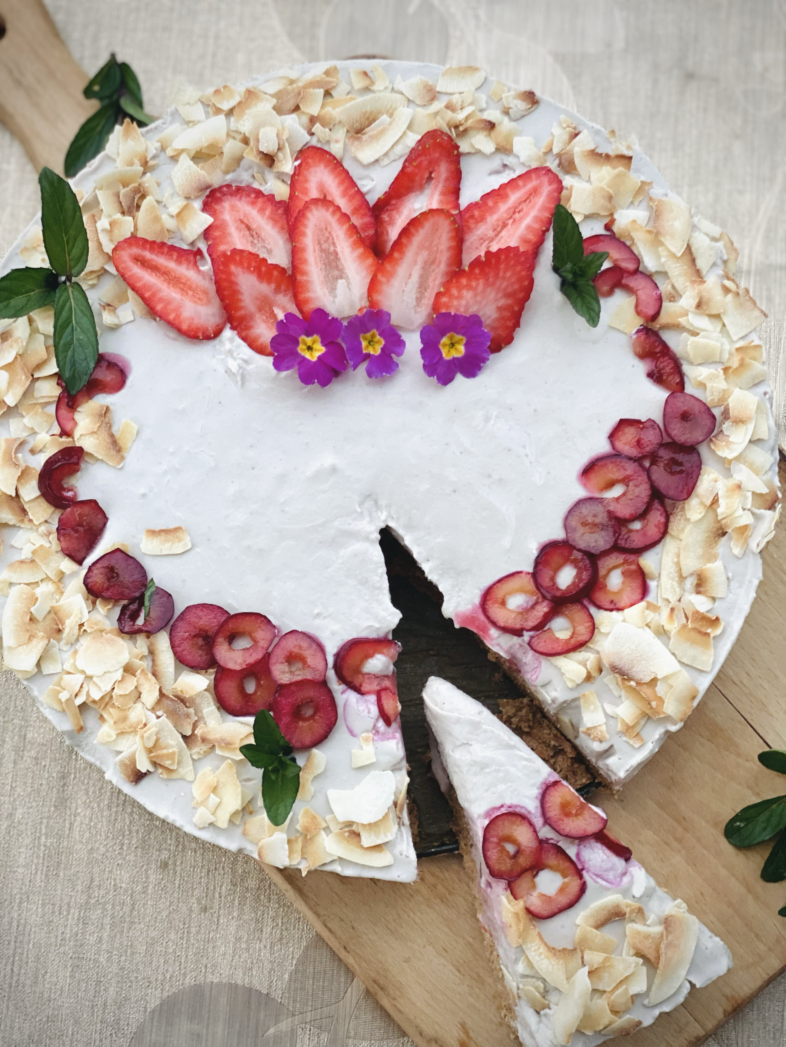 A round white frosted cake decorated with sliced strawberries, red grapes, white chocolate shavings, purple flowers, and green leaves on top. One slice is cut and slightly removed, revealing a chocolate layer inside.