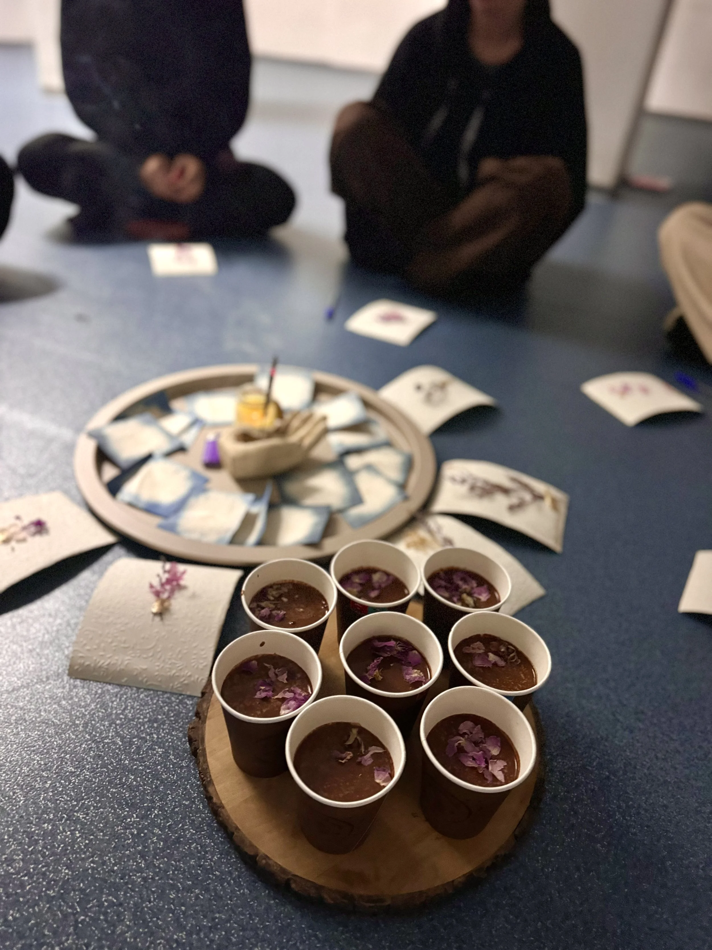 Several small cups of tea with purple flower petals on top arranged on a wooden round tray on a table, surrounded by papers and other objects, with two people sitting in the background.