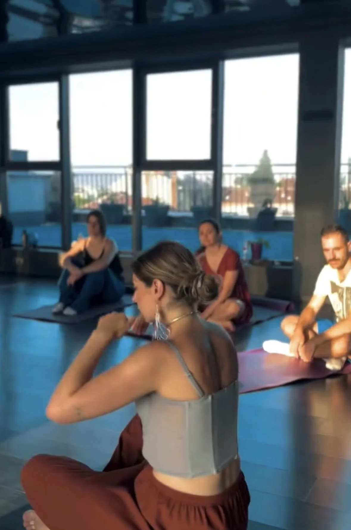 People participating in a yoga or meditation class in a bright studio with large windows showing an urban skyline.