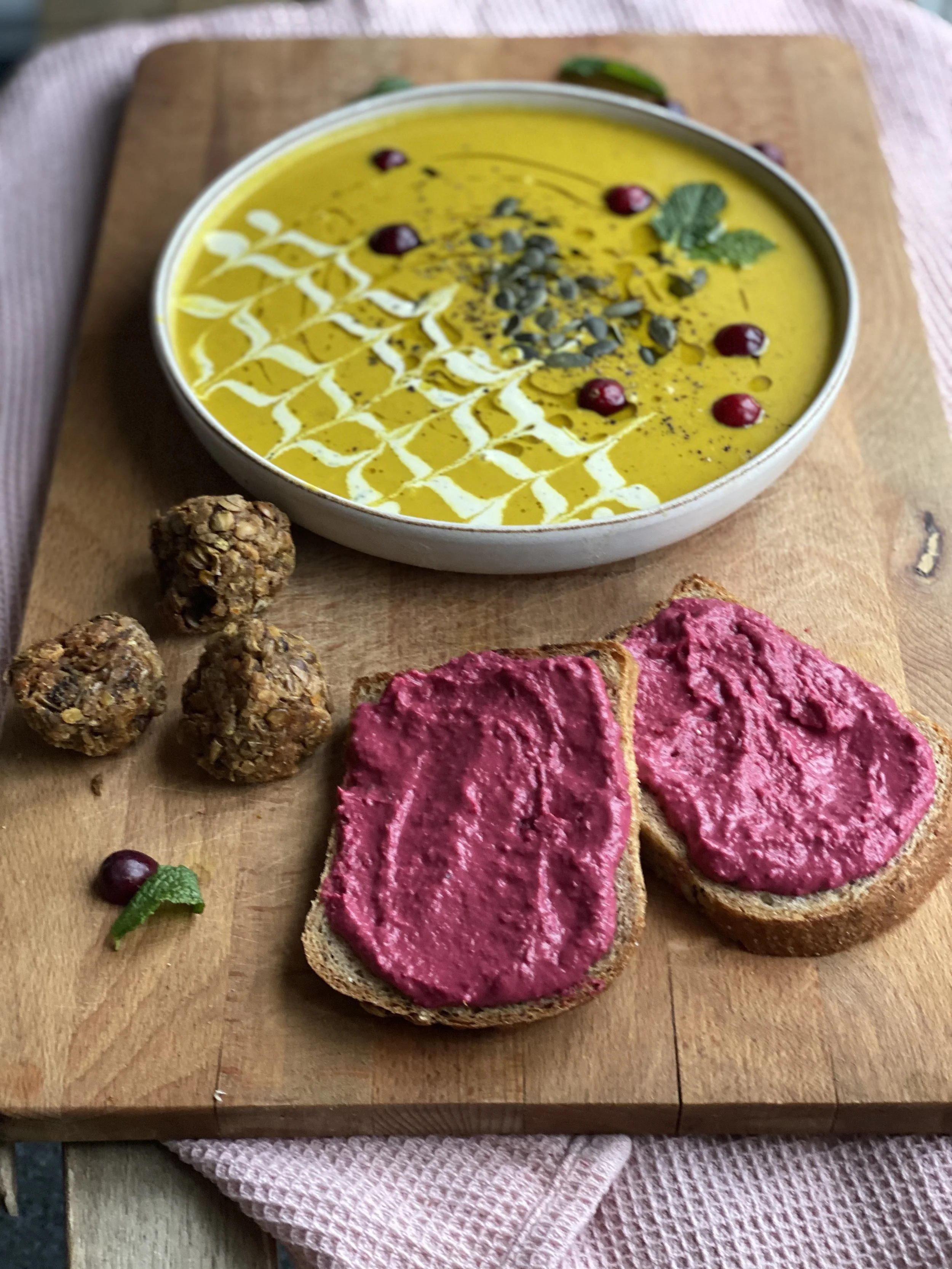 A bowl of yellow soup garnished with cream drizzle, red berries, seeds, and mint leaves on a wooden board. Next to it are two slices of bread with pink spread, three falafel balls, a mint leaf, and a red berry.