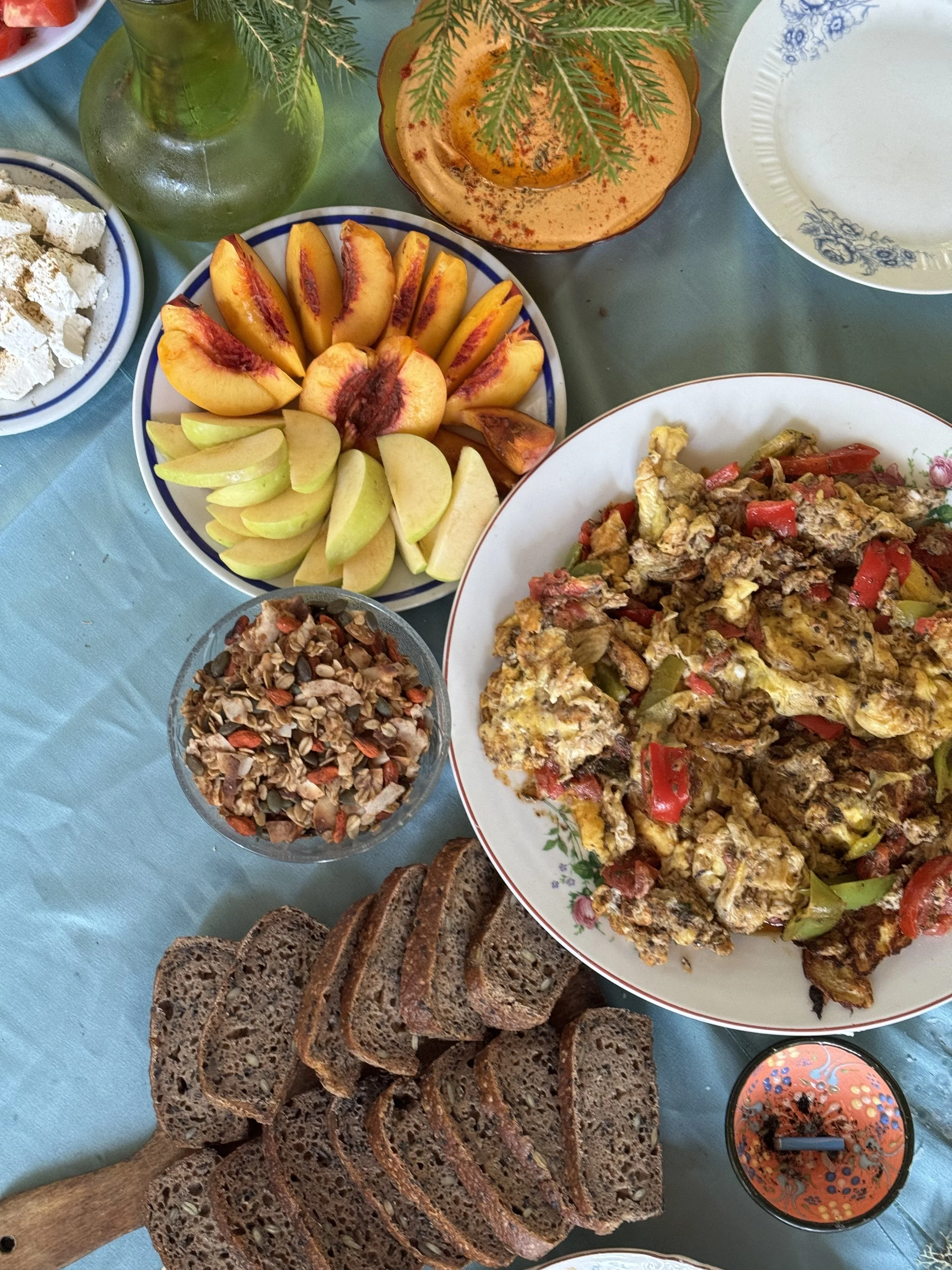 A spread of food on a table including sliced bread, a large plate of scrambled eggs with vegetables, a bowl of granola, a plate of assorted fresh sliced peaches and apples, a glass dish of salad or dip, a bottle of olive oil or vinegar with green her