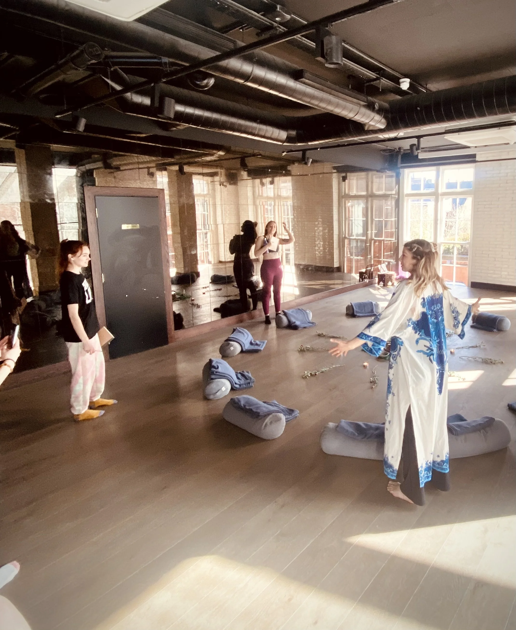 A woman dressed in a white and blue robe is leading a yoga or stretching class in a studio with natural light. There are yoga mats, towels, and props on the floor, and a large mirror on the wall reflecting the participants. Several people, including 