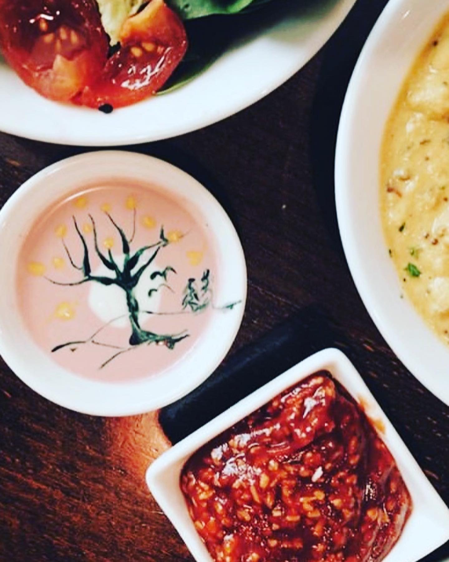 Assorted dishes including a salad with cherry tomatoes, a dish with a yellow sauce or soup, a bowl of a pink beverage with a drawing of a tree and birds, and a small bowl of spicy red chili sauce.