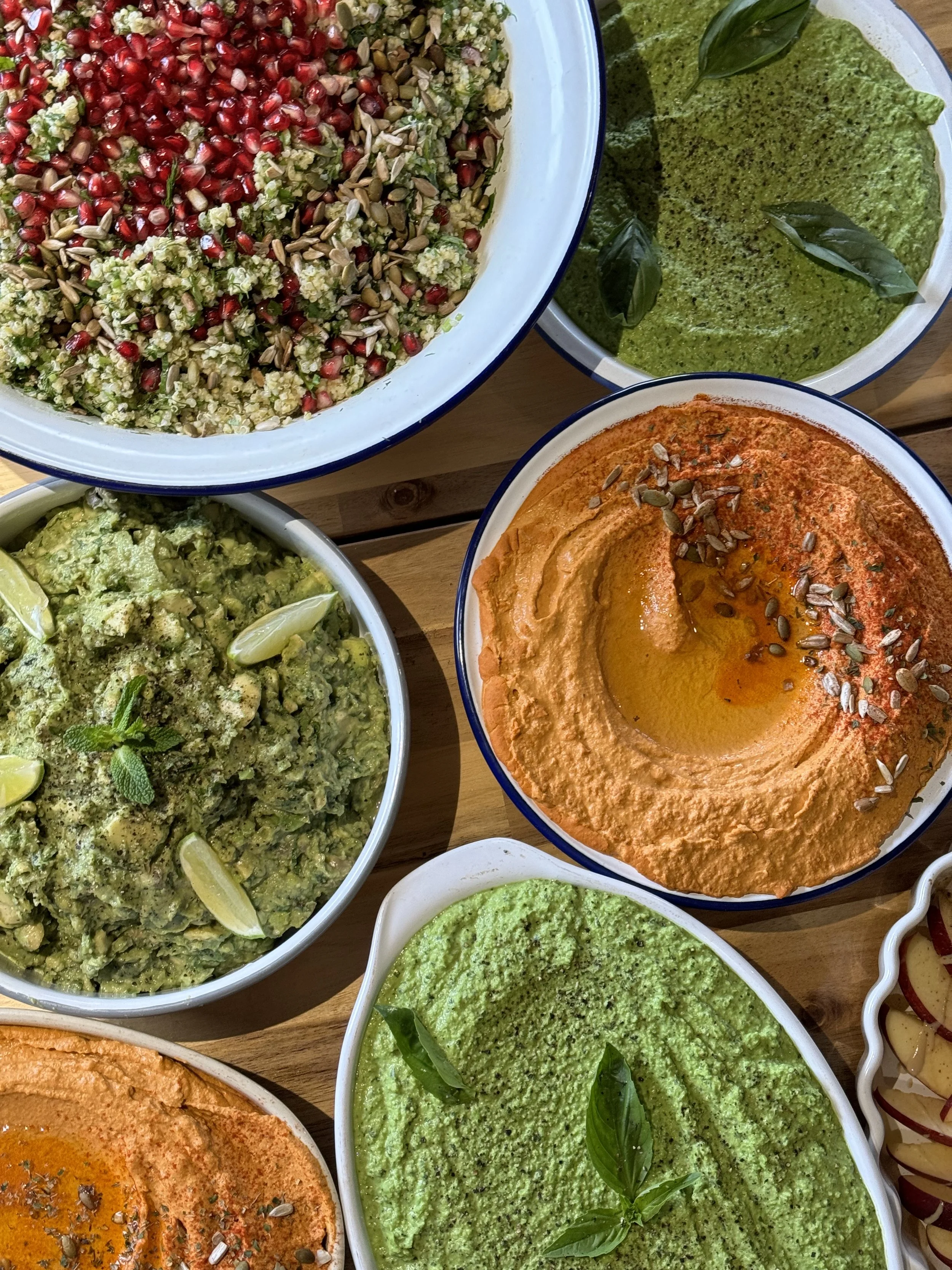 An assortment of bowls containing various dips and spreads, including hummus, guacamole, green pesto, and a salad with pomegranate seeds, sunflower seeds, and chopped vegetables.