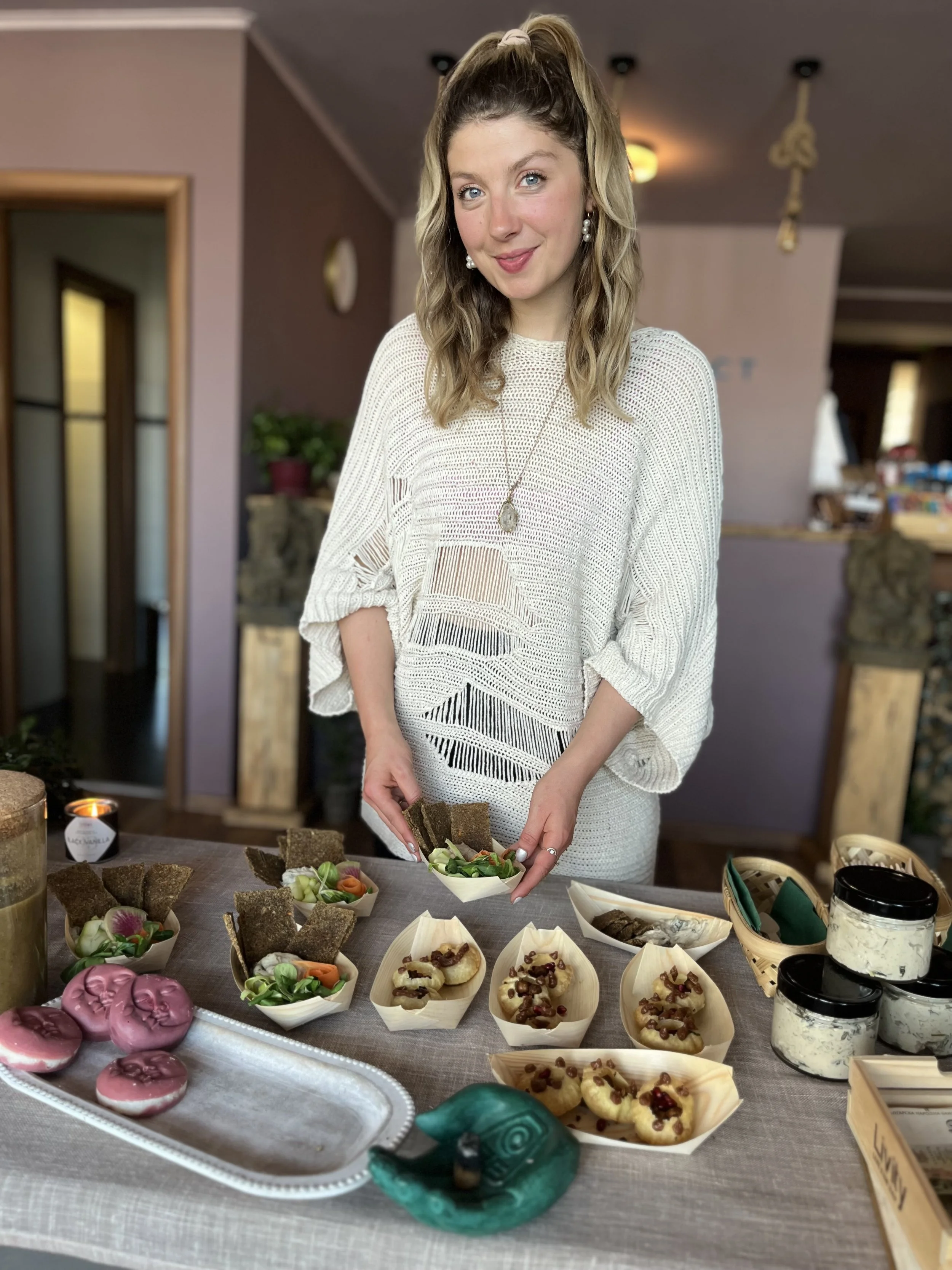 A woman with long wavy blonde hair, wearing a white knitted sweater and jewelry, standing behind a table with various food items, including small bowls of salad, sandwiches, cookies, and jars of dip, in a cozy indoor setting.