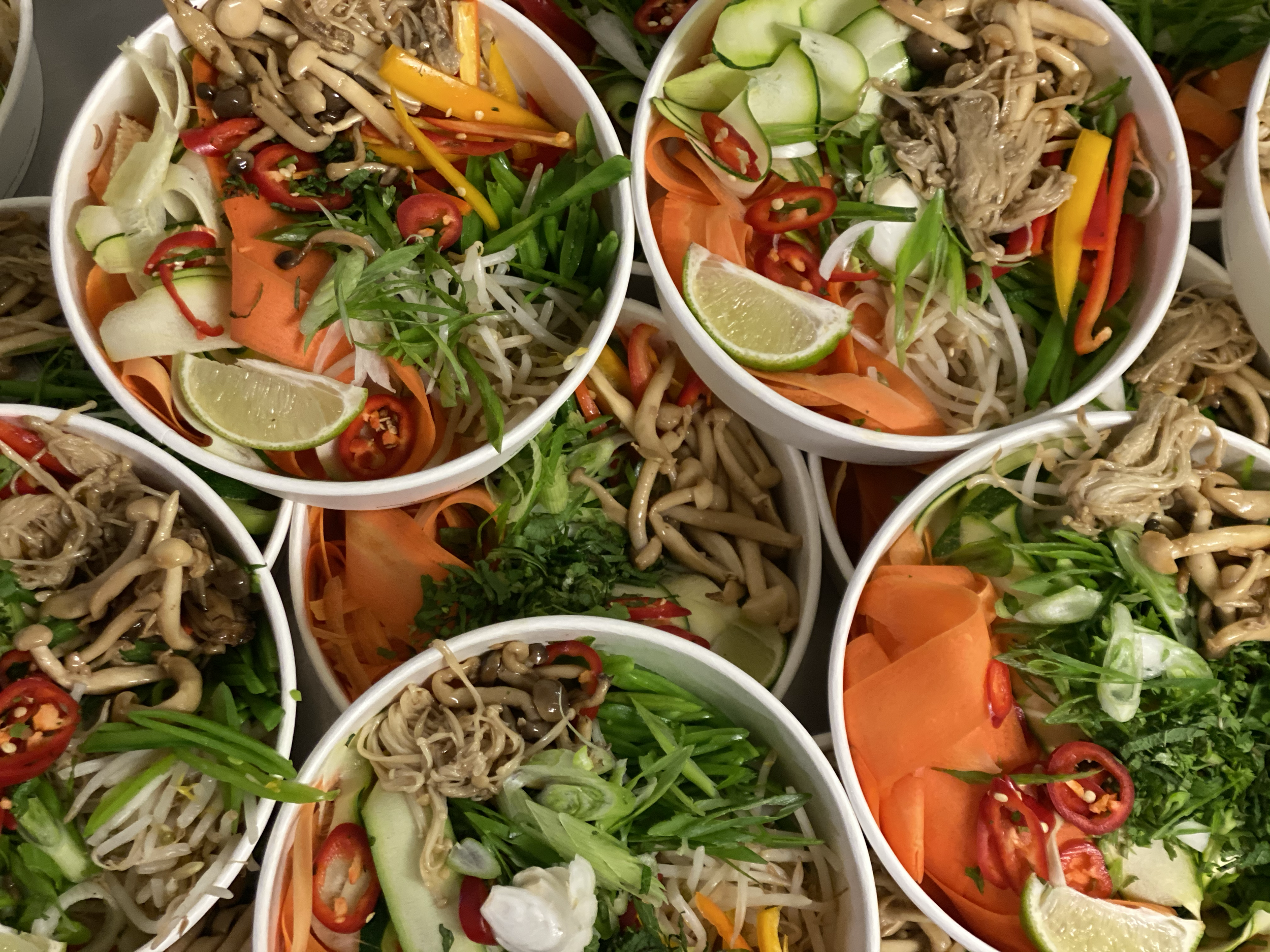 Multiple bowls of fresh Asian-style noodle salad with vegetables, herbs, sliced chili peppers, and lime wedges.