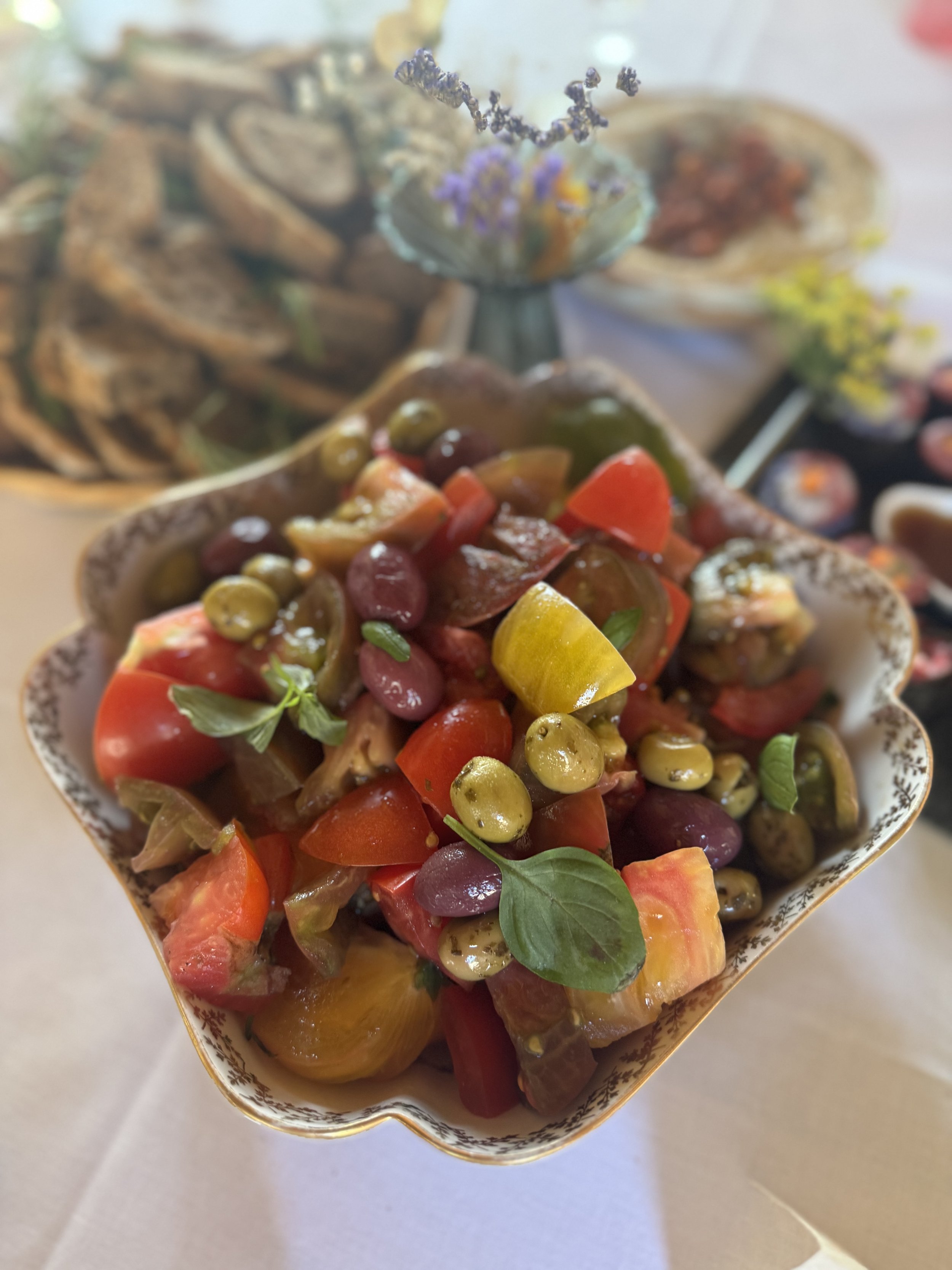 Close-up of a bowl of colorful mixed fruit salad with grapes, tomatoes, and herbs, on a white table with a blurred background of plates and a flower vase.