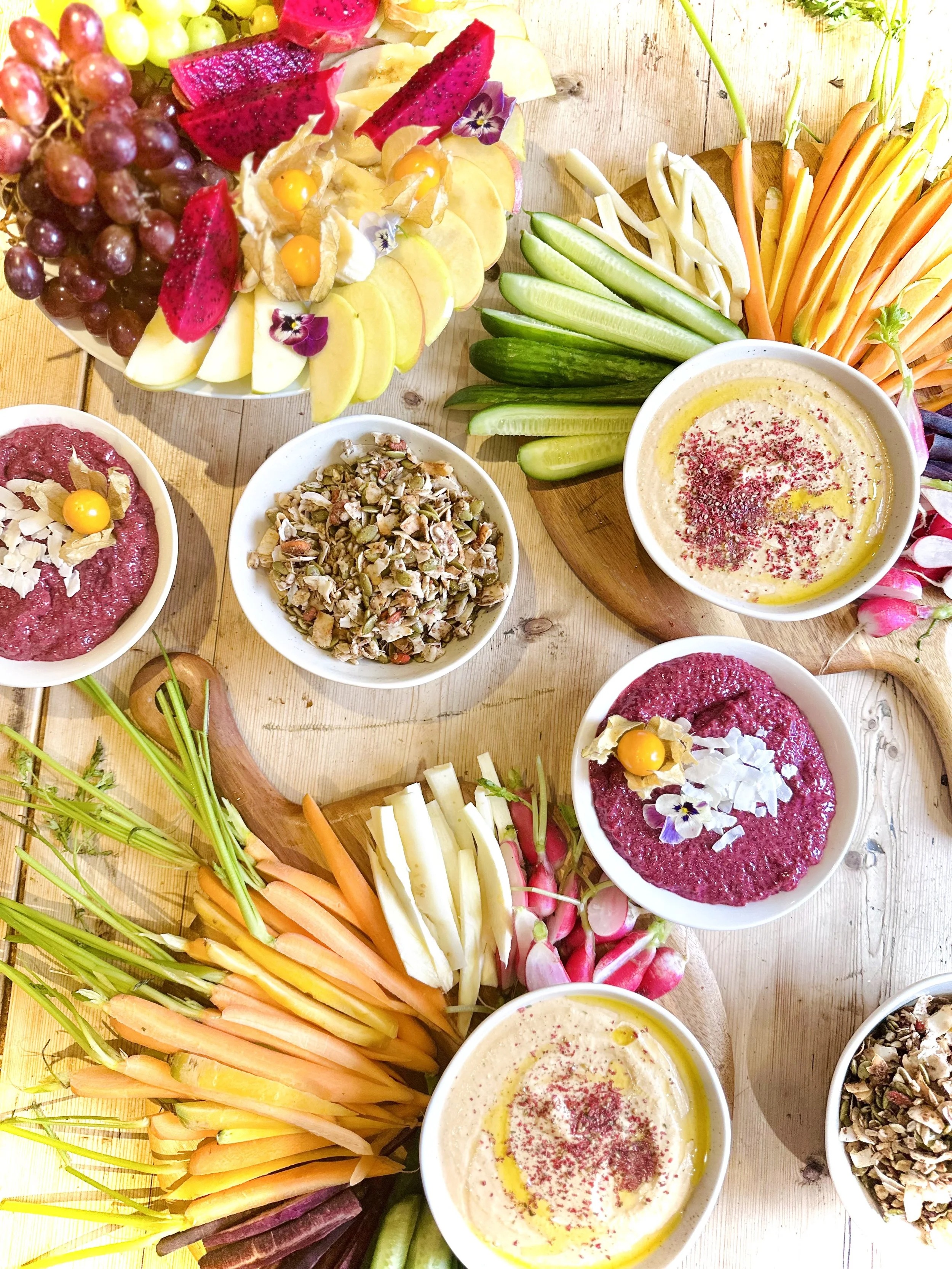 An assortment of fresh fruit, vegetables, dips, and spreads on wooden tables, including grapes, sliced fruits, carrots, celery, radishes, hummus, and other small bowls of food, decorated with edible flowers.