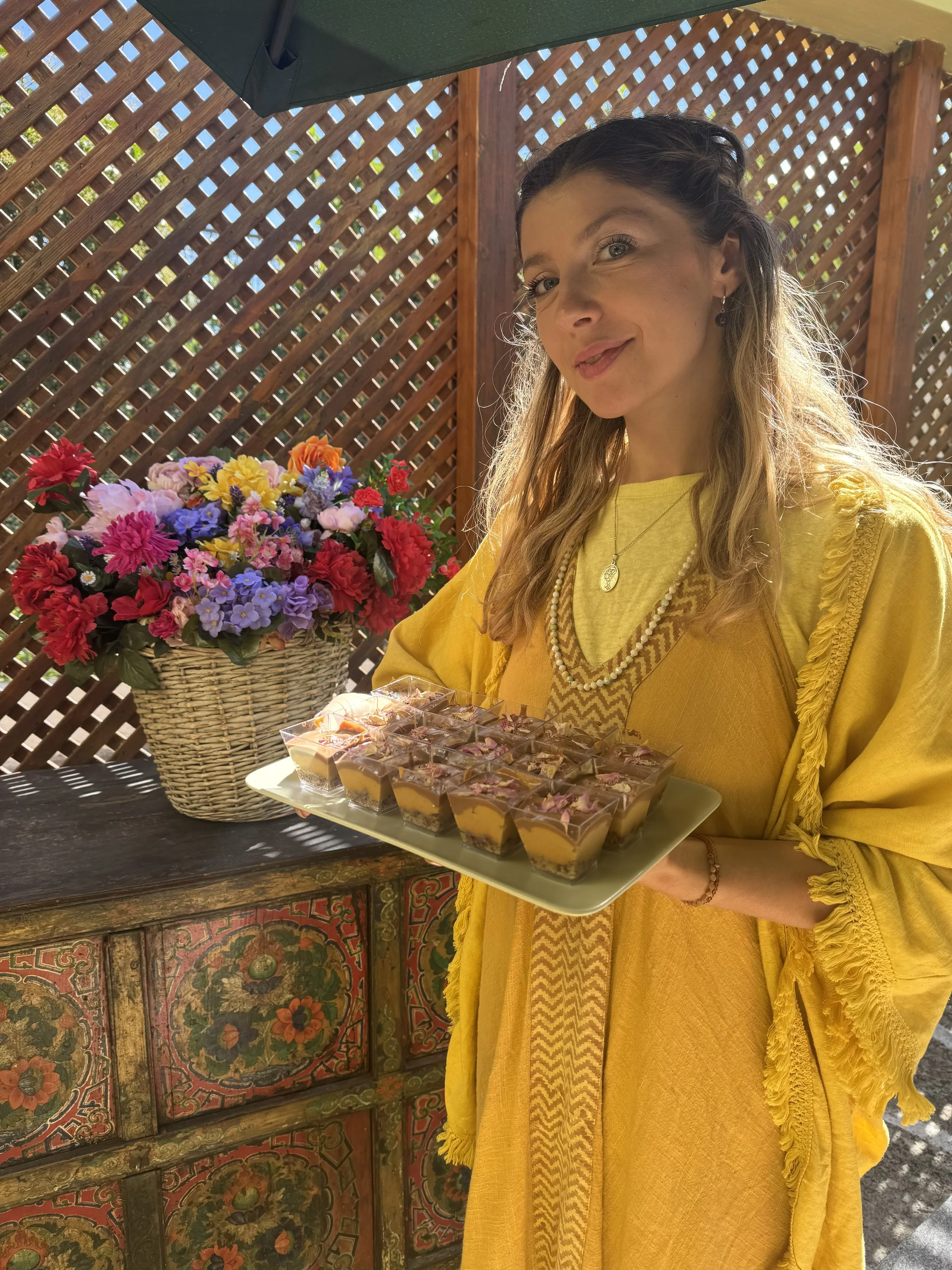 A woman wearing a yellow dress and jewelry holding a tray of small dessert cups with pink and purple toppings, standing next to a large basket of colorful artificial flowers, against a wooden lattice backdrop under a green umbrella.