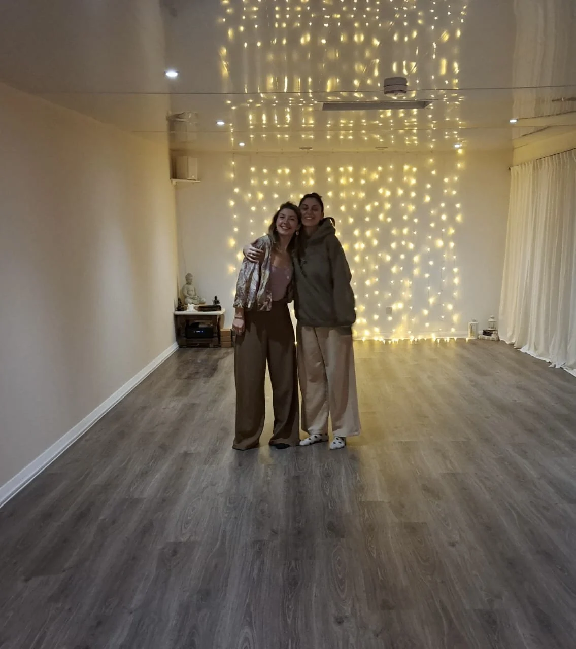 Two women standing arm in arm in a room with wooden flooring and string lights on the wall behind them, smiling at the camera.