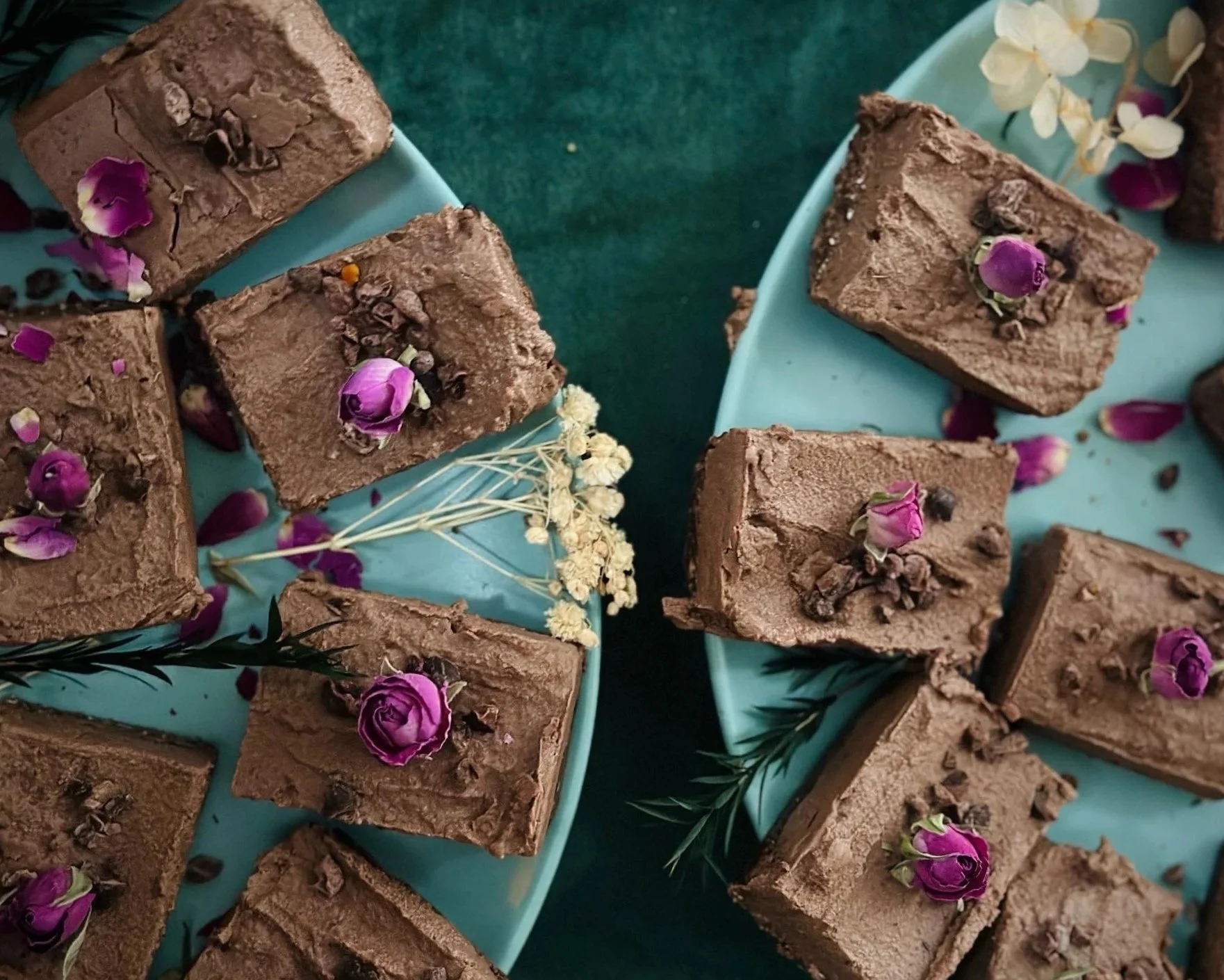 Several squares of chocolate fudge topped with dried rose petals, small chocolate chips, and herbs on turquoise plates with a green background.