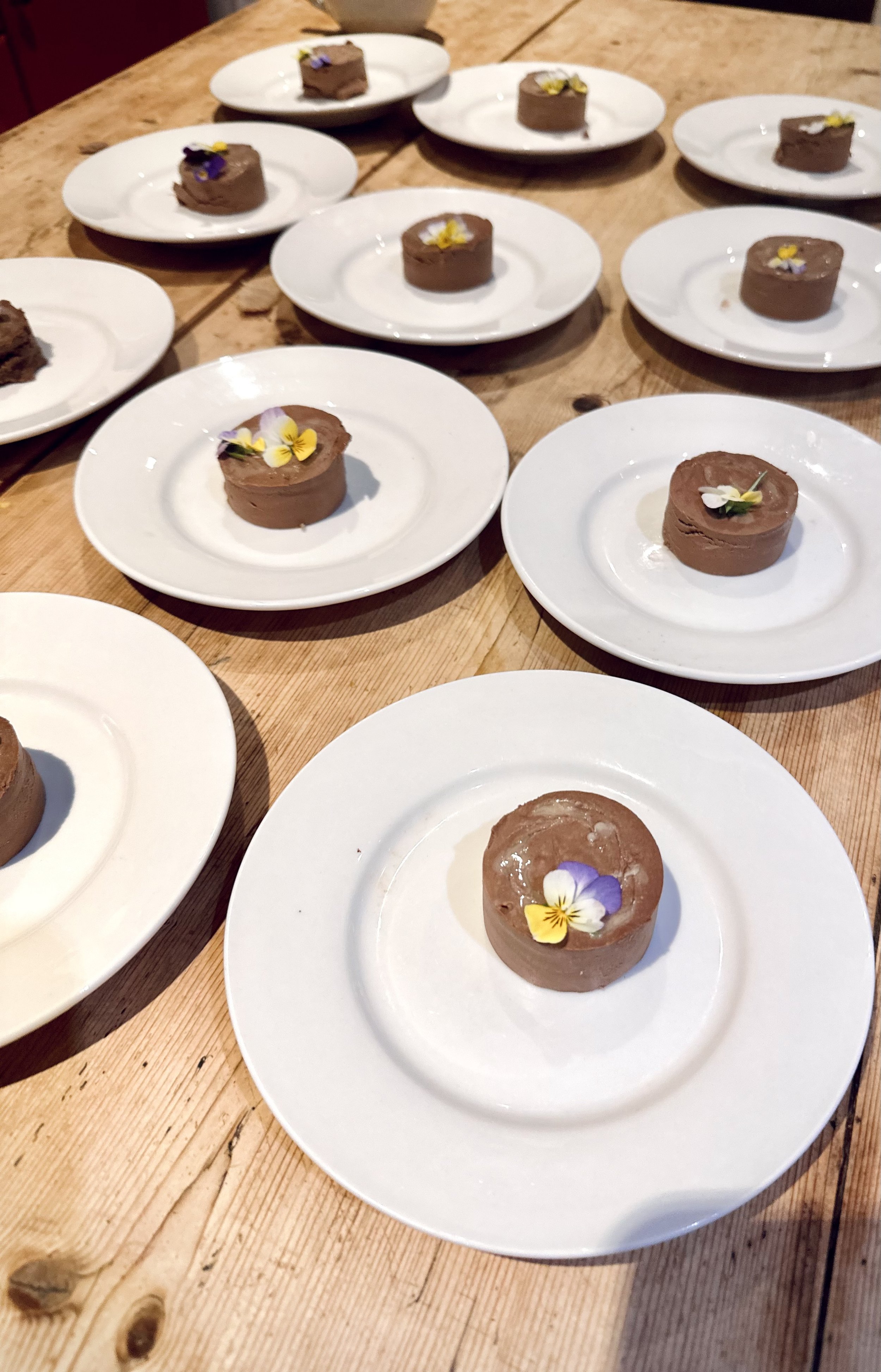 Multiple small chocolate ice cream desserts with edible flowers on white plates on a wooden table.