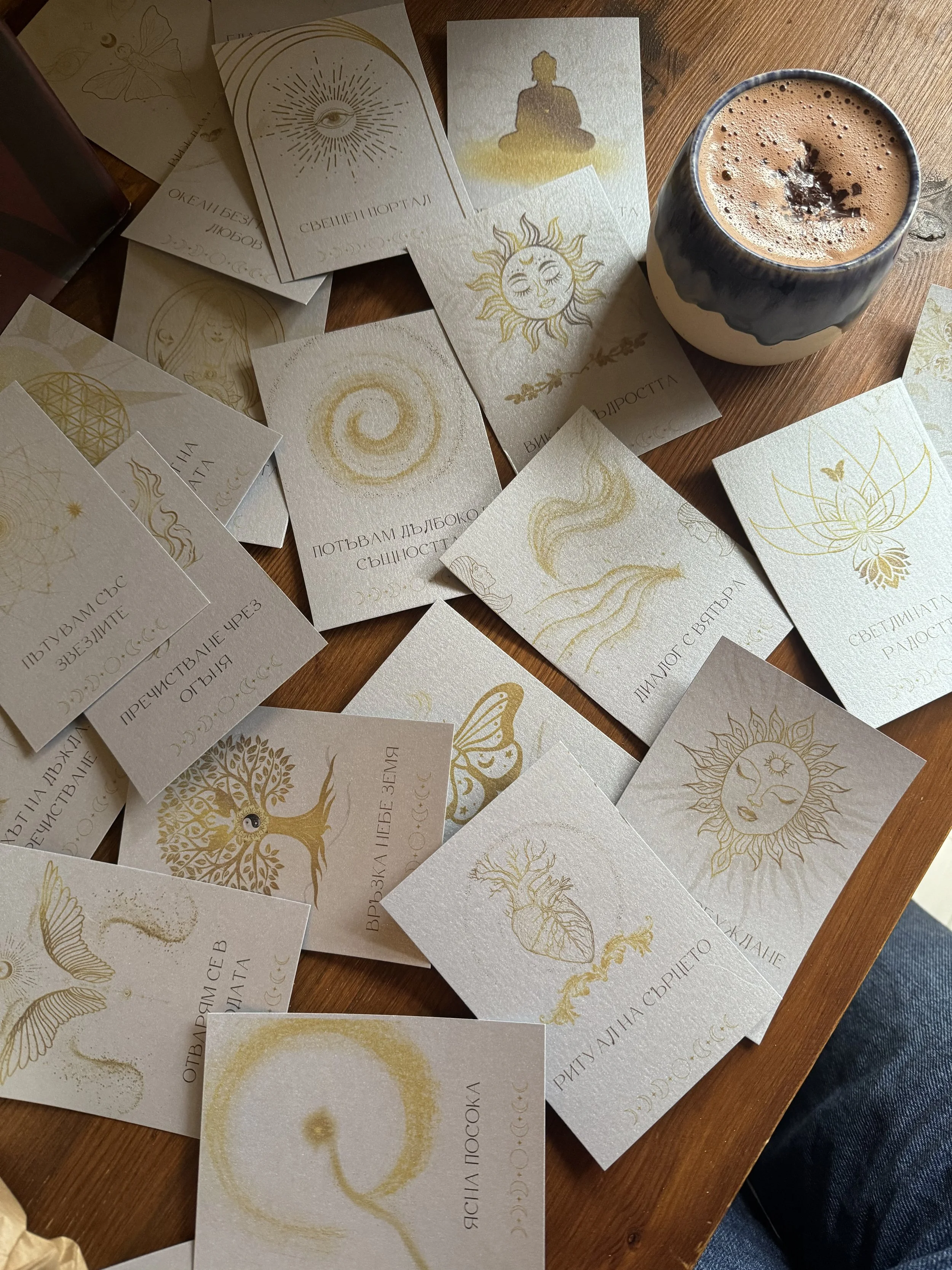 Various oracle and tarot cards with gold illustrations and Cyrillic text spread out on a wooden surface, alongside a cup of hot chocolate or coffee in a black and white mug.