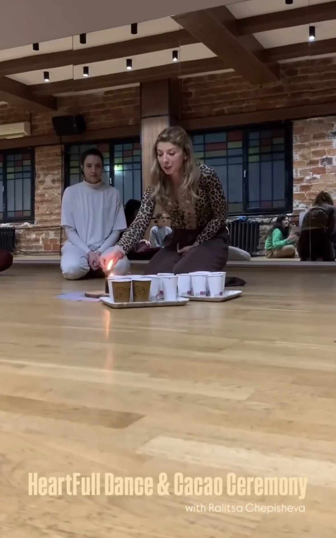 A woman performing a cacao ceremony indoors, with a lit candle and cups on a tray, while a woman in white sits nearby, and others observe in the background.