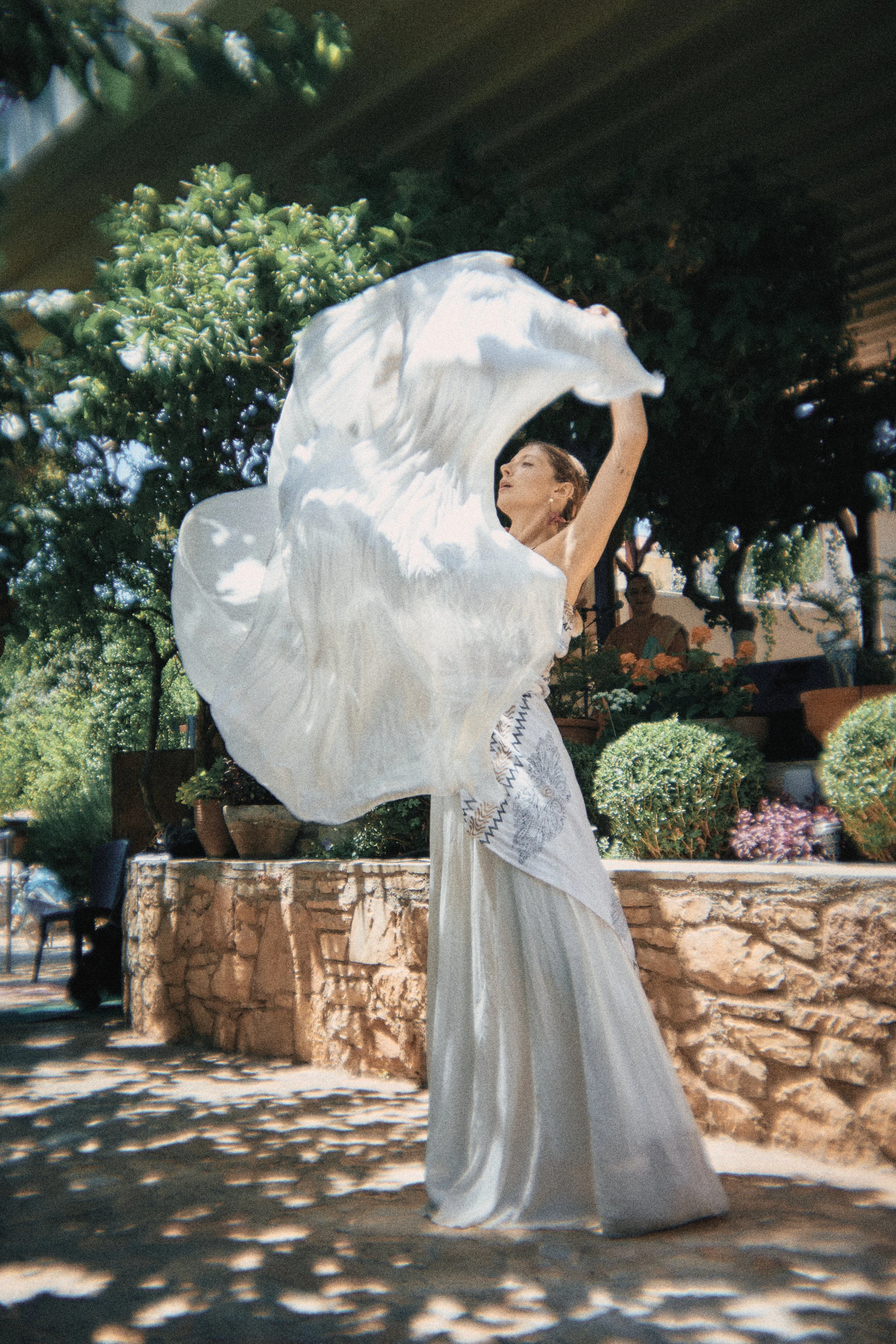 A woman in a long, elegant white dress dances outdoors, holding a flowing white fabric above her head. She is surrounded by plants and potted greenery on a sunny day.