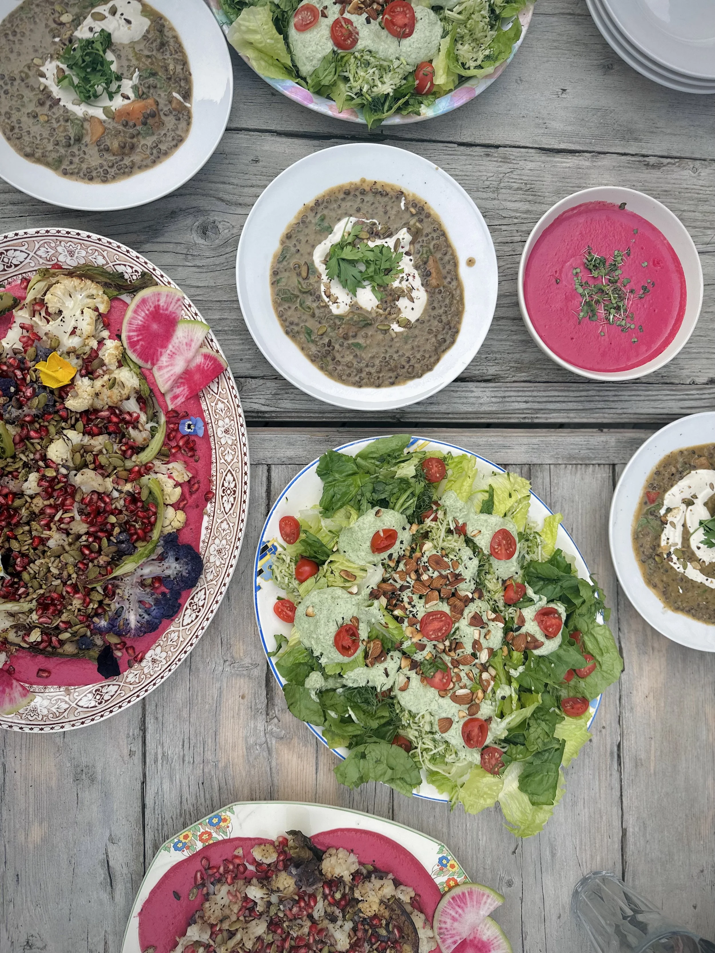 Assorted salads and bowls of soup on a rustic wooden table, including a green salad with cherry tomatoes, a pink beet-based dish, and a dish with lentils and vegetables.