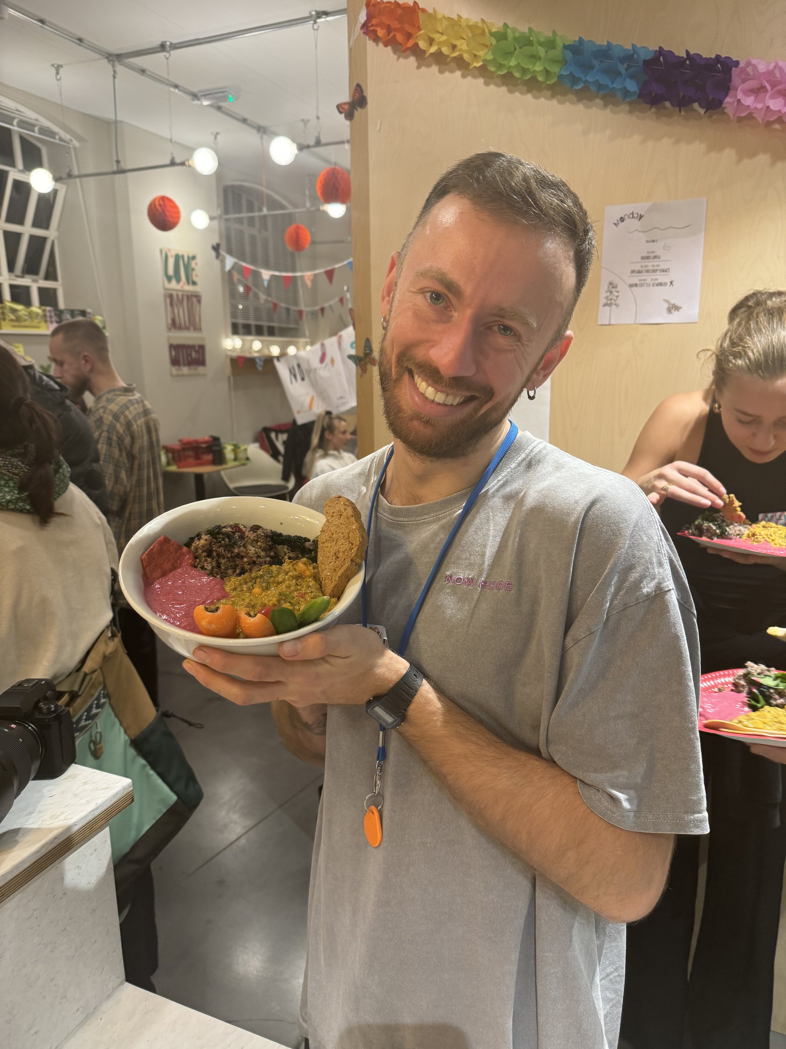A young man with short dark hair, beard, and earrings smiling and holding a bowl filled with colorful food, standing in a decorated indoor setting with birthday banners, hanging paper lanterns, and other people in the background.