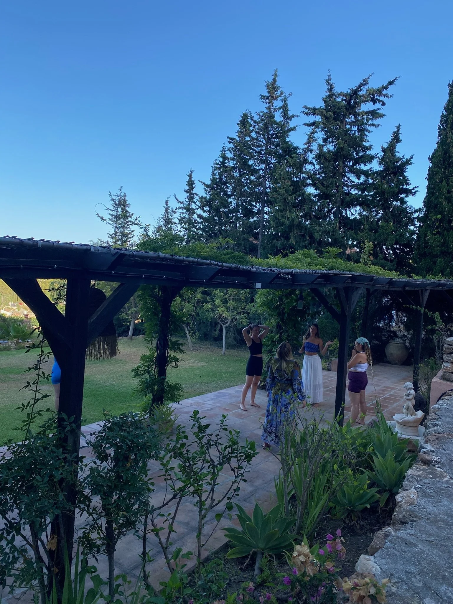 Four women standing and conversing on a tiled patio under a dark pergola, with a garden and tall trees in the background on a clear evening.