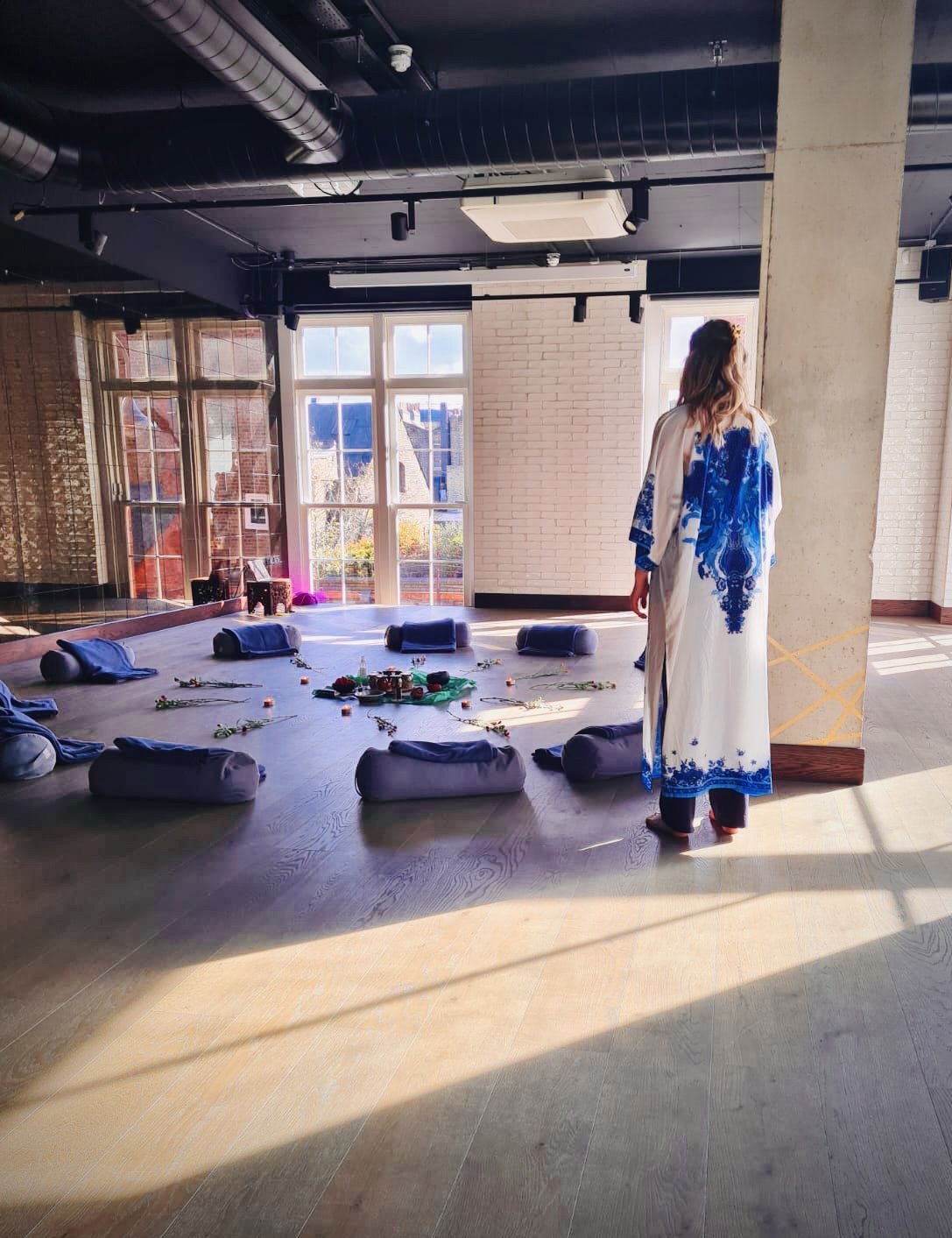 A woman in a white and blue garment standing in a room with sunlight streaming in, with cushions and decorative items arranged on the floor in a circular pattern, windows showing a sunny outdoor scene, and an industrial-style ceiling.