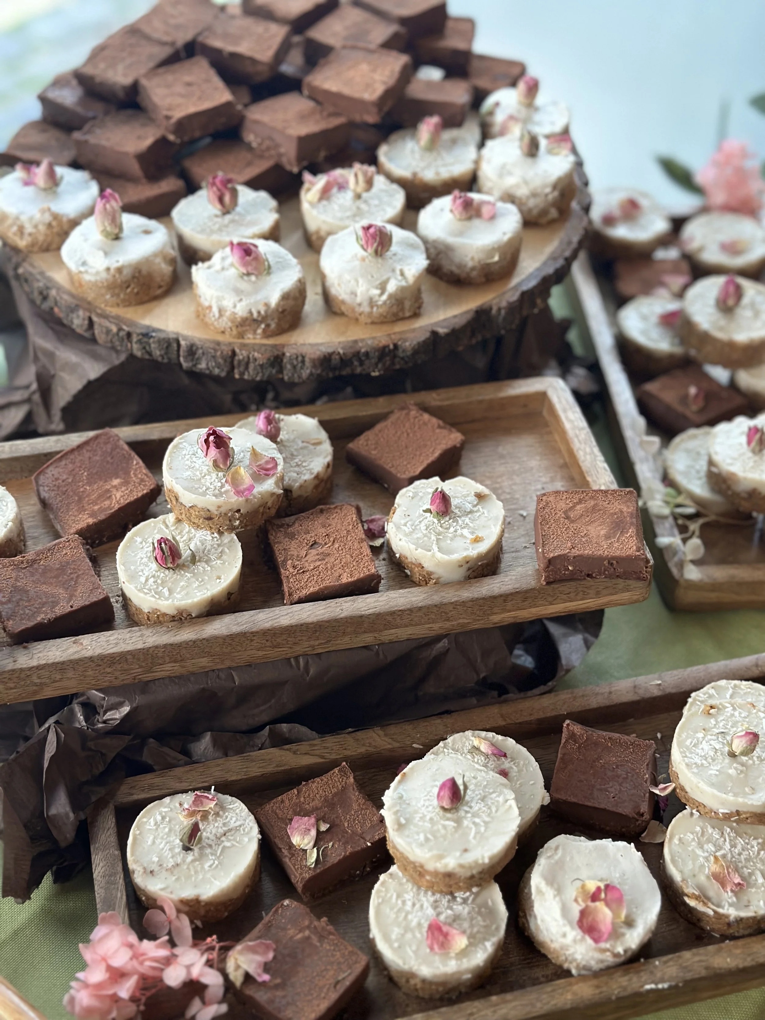 Assorted dessert bites on wooden trays, including cheesecakes topped with pink rose petals and chocolate brownies, arranged on rustic wooden platforms.