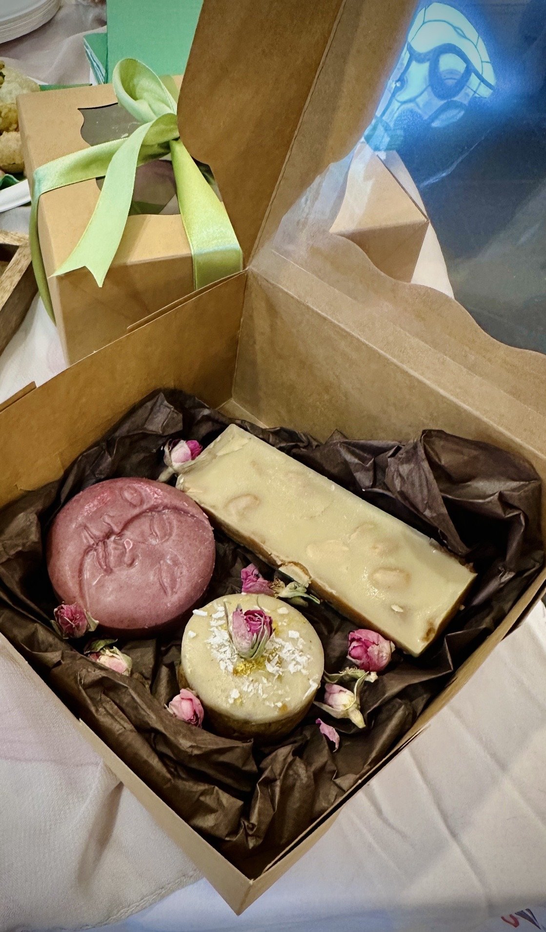 A gift box contains two round soaps and a rectangular soap bar, decorated with pink rosebuds and white flower petals, surrounded by crumpled brown paper.