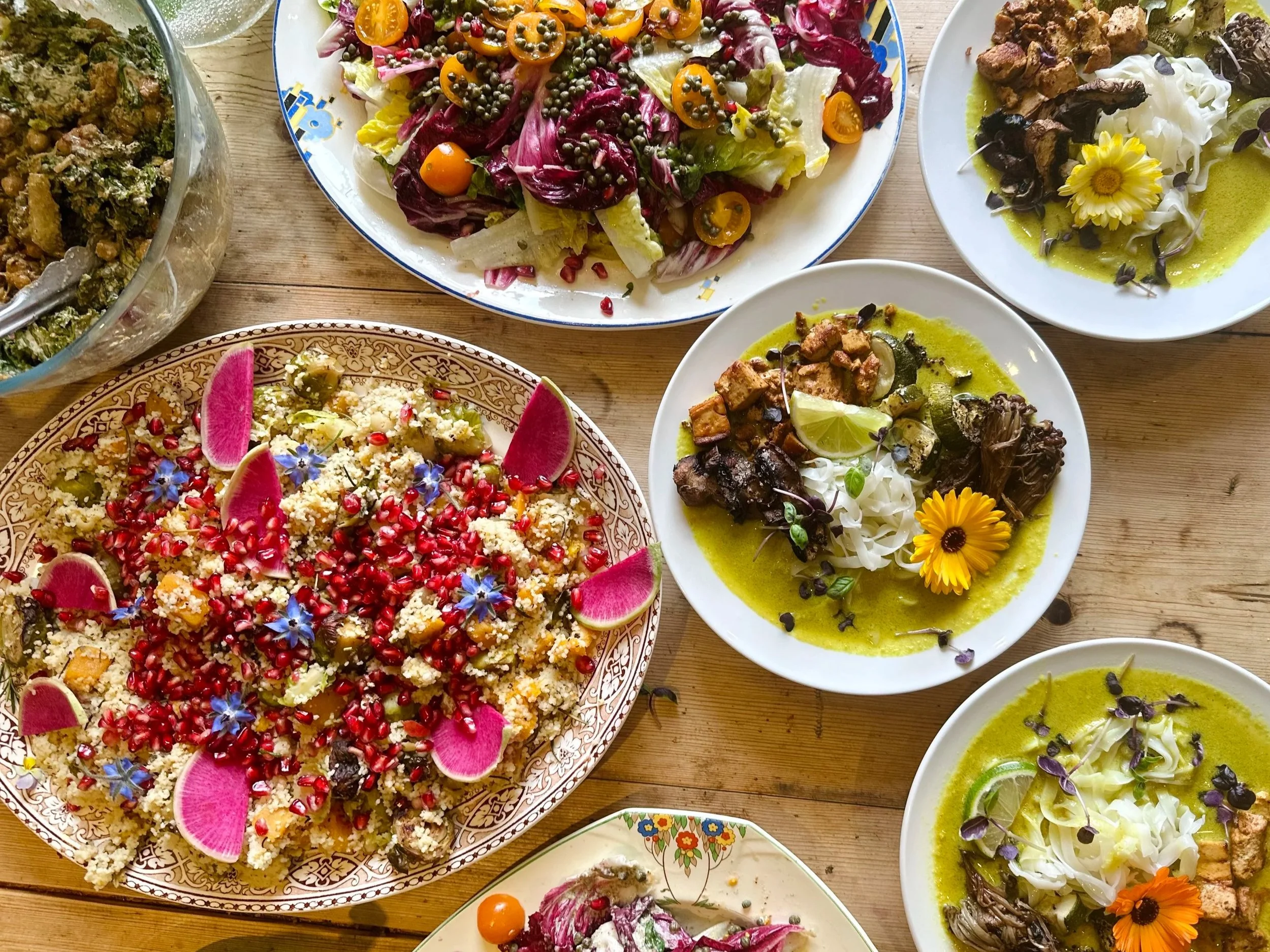 A variety of colorful dishes including salads, grains, and soups on a wooden table, garnished with flowers, seeds, and fruits.