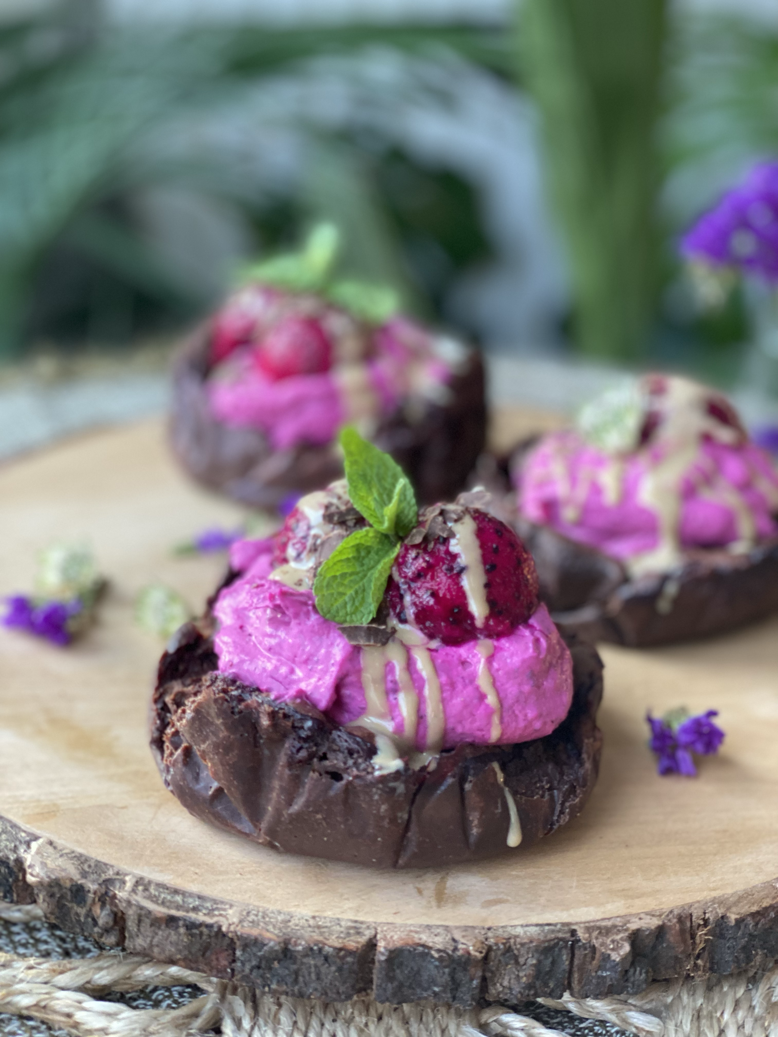 Chocolate dessert topped with pink cream, strawberries, chocolate shavings, and mint leaves on a wooden surface with flowers in the background.