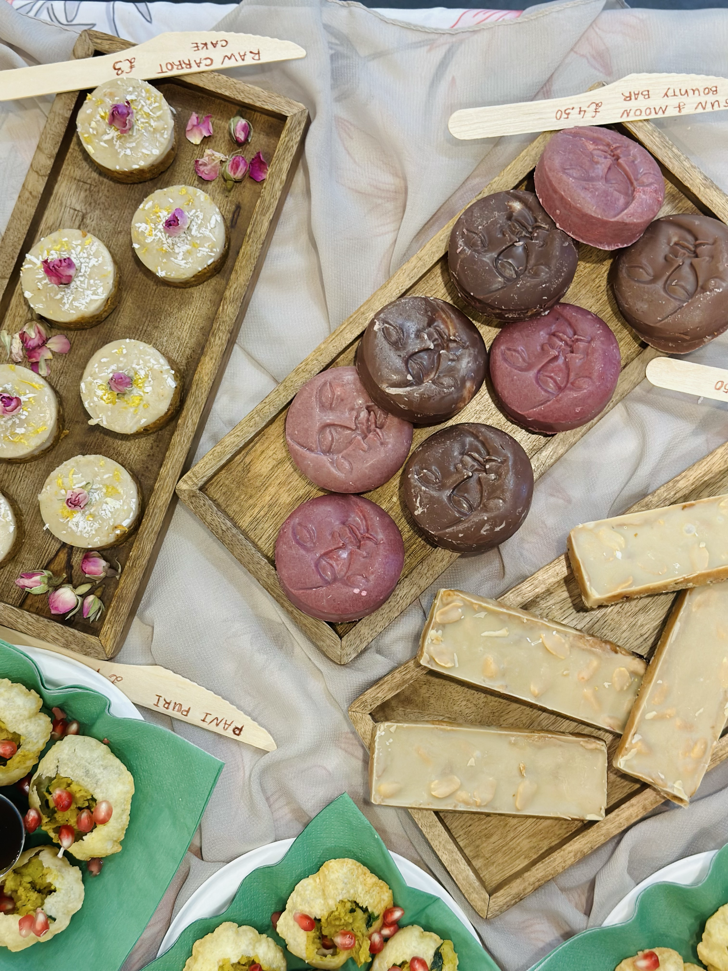 Display of assorted desserts including coconut-covered treats with pink rose petals, round chocolate and pink confections with embossed faces, nut-topped white chocolate bars, and green-frosted sweet bites with pomegranate seeds, all on wooden and ce