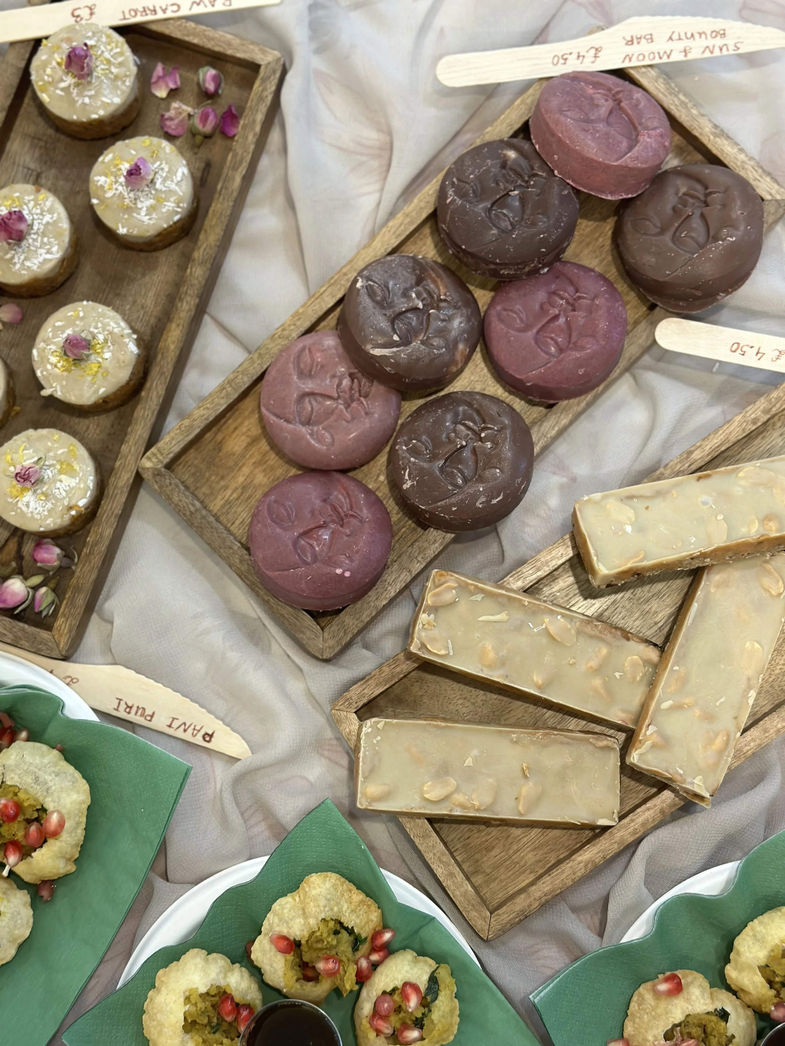 Assorted baked goods on wooden trays, including round cookies with glazes and floral decorations, round chocolate and berry-colored candies with embossed faces, and rectangular almond-topped white chocolate bars, placed on a light fabric surface.