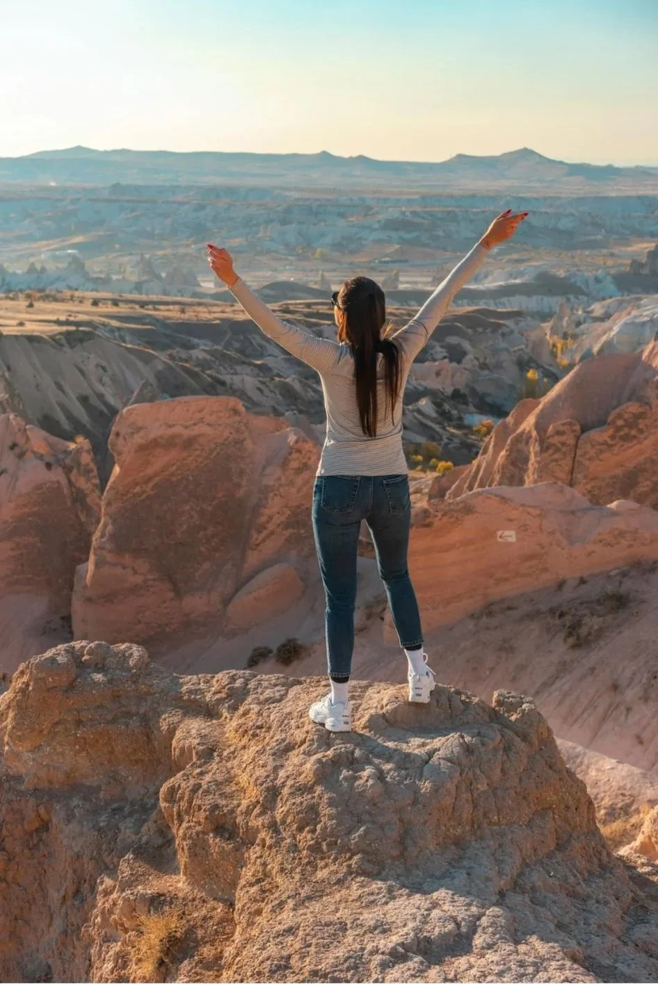 A woman stands on a large rock with arms raised, overlooking a canyon landscape with colorful rock formations and distant hills.