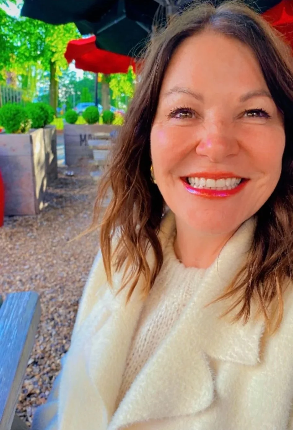 Smiling woman with brown wavy hair wearing a beige sweater sitting outdoors at a table under a red and black umbrella, with trees and green bushes in the background.