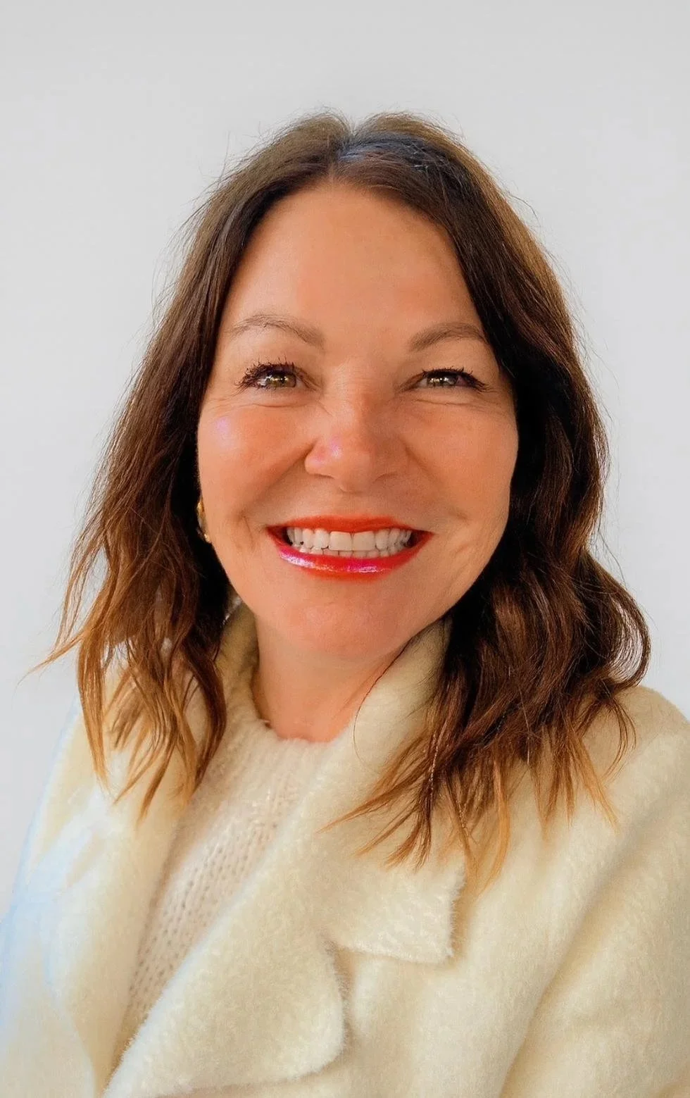 A woman with shoulder-length brown hair smiling broadly, wearing a cream-colored sweater, against a plain white background.