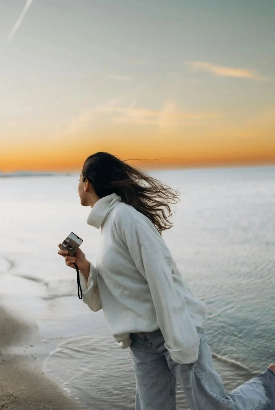 A woman with windblown hair standing on the beach at sunset, holding a camera in her hand.