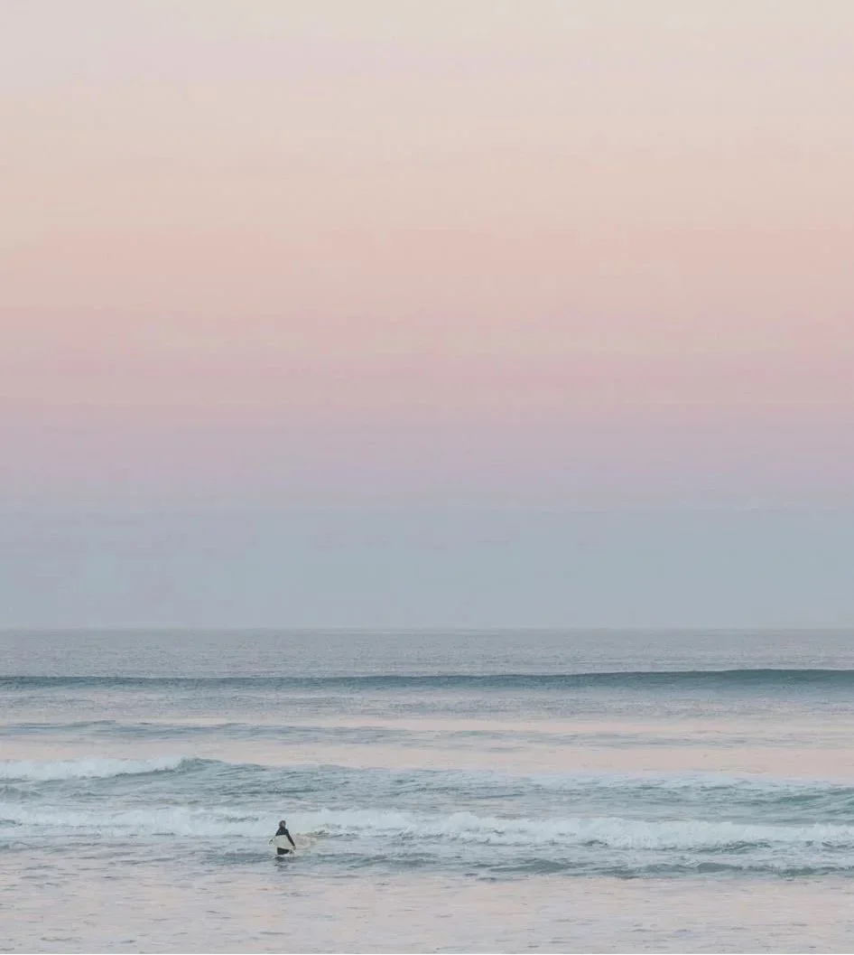 A person standing in the ocean waves during sunset or sunrise with a pastel-colored sky.