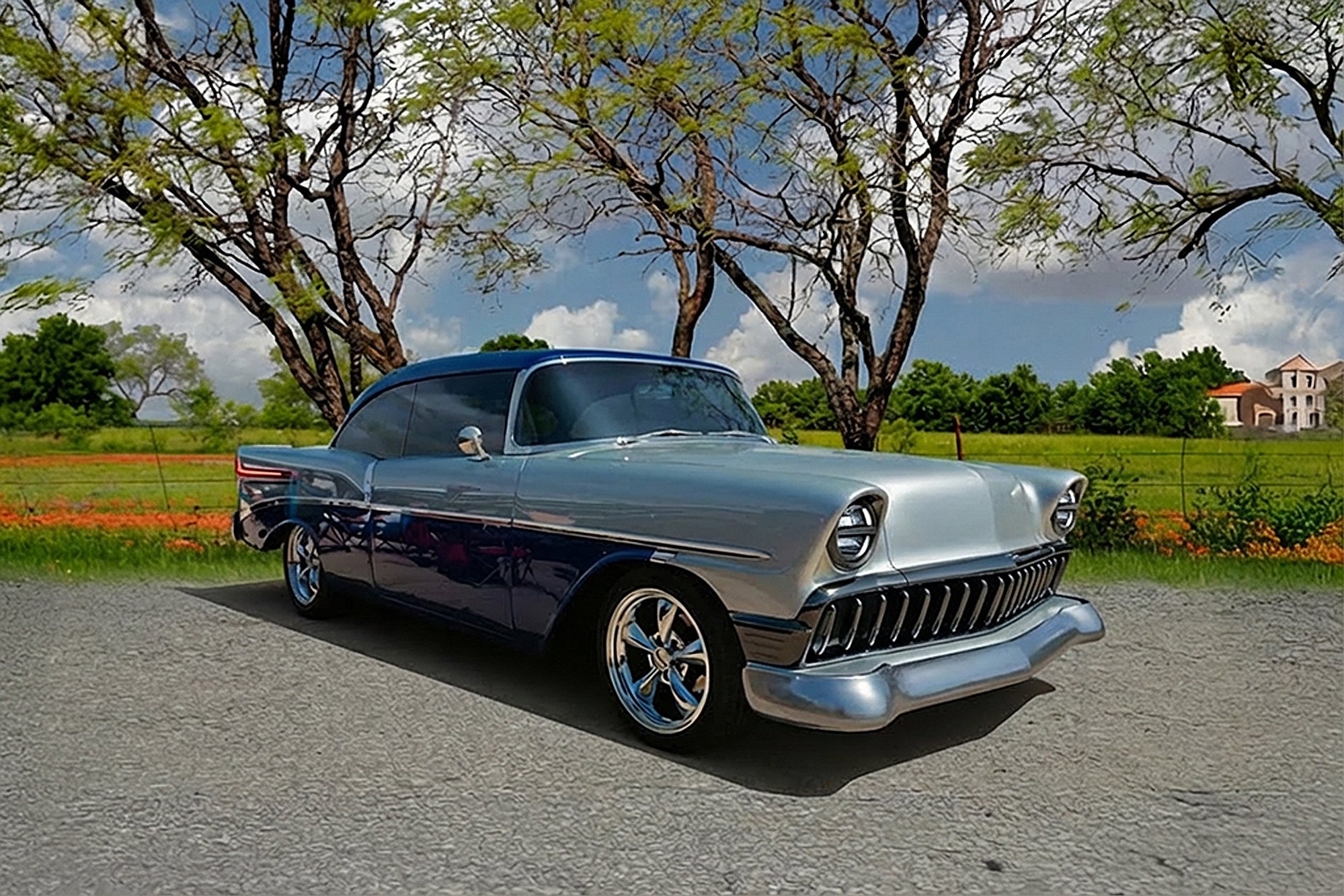 A classic silver and black vintage car parked on a gravel road, with leafless trees and a blue sky with clouds in the background.