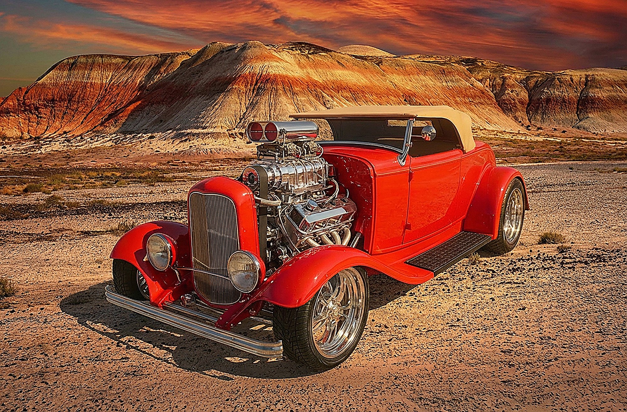 A vintage red car with a large engine and chrome details parked on a desert landscape at sunset, with colorful clouds and mountainous terrain in the background.