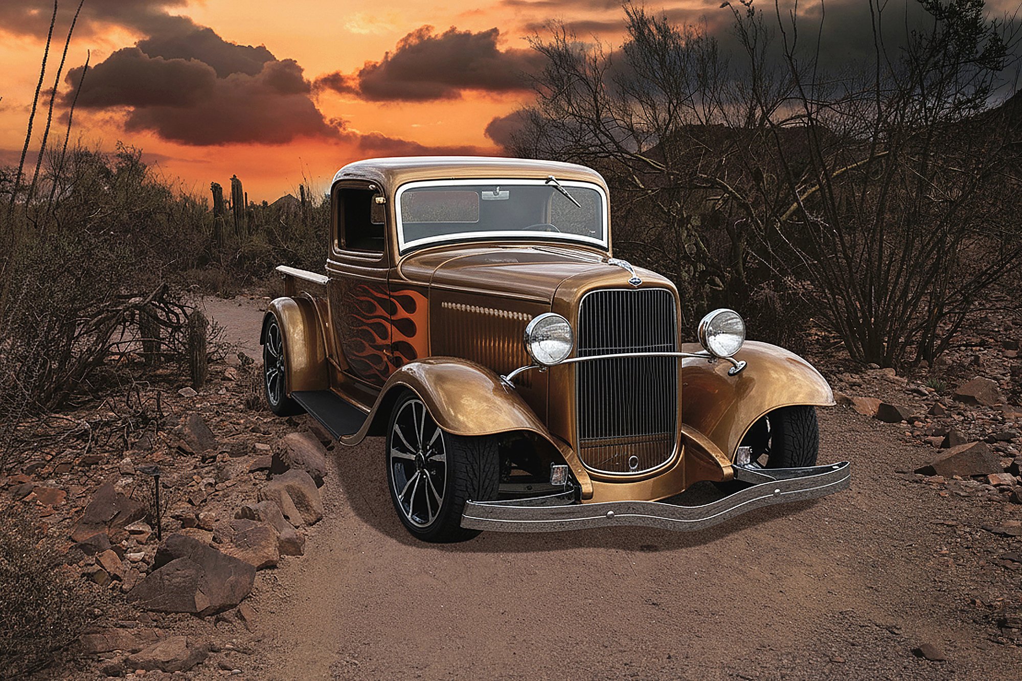 A vintage brown pickup truck with flame decals on the side parked on a desert dirt road at sunset, with dry bushes and cacti around and dark clouds in the sky.