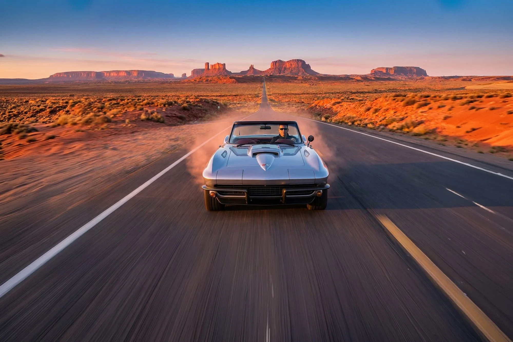 A vintage silver sports car with a driver wearing sunglasses driving on an empty desert highway at sunset, with red rock formations in the background.