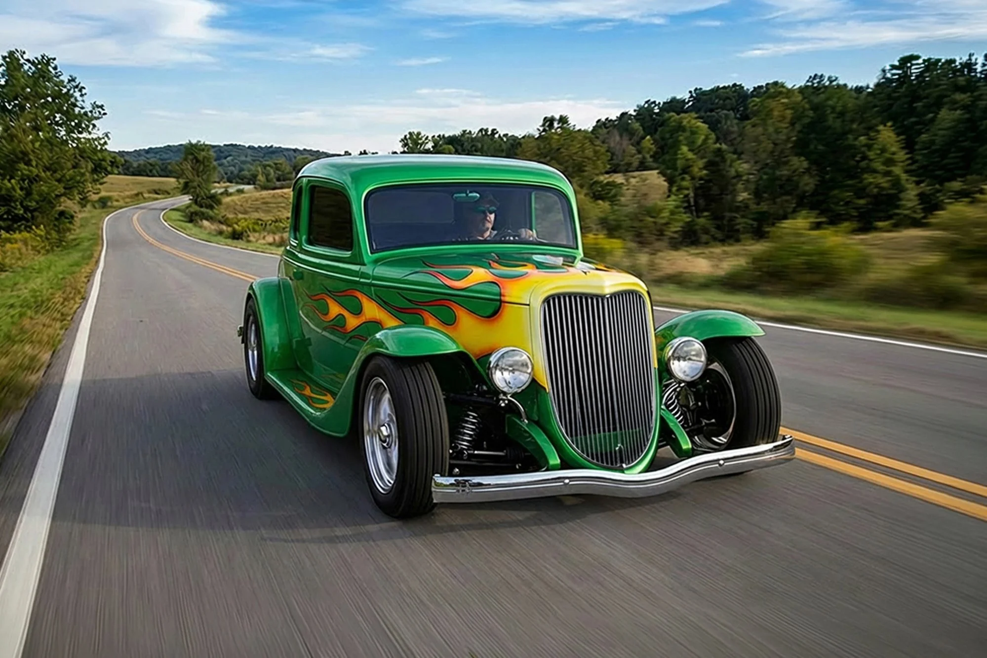 A vintage green car with flame decals driving on a winding country road surrounded by fields and trees under a partly cloudy sky.