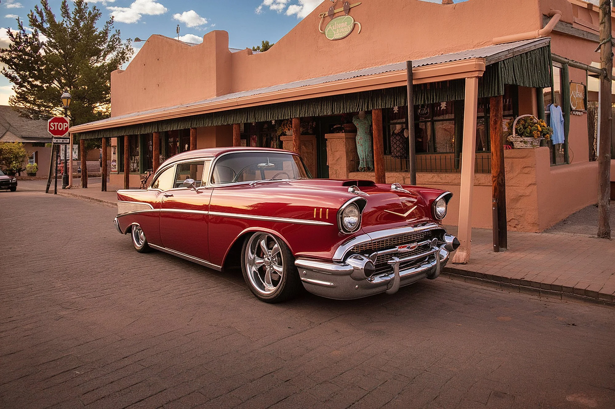 A vintage red and white classic car parked on a brick street in front of a pink building with a green awning, a mannequin in the window, and a basket of flowers. A stop sign and a tree are visible in the background.