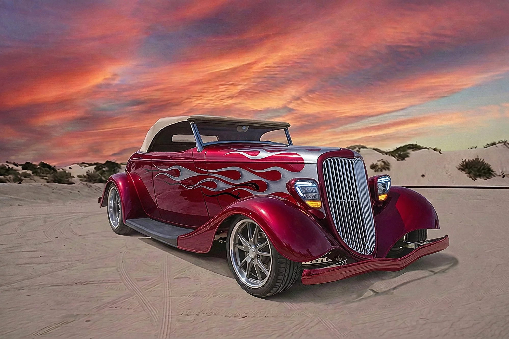 A vintage red car with flame decals parked on a sandy desert terrain during a colorful sunset with pink, orange, and purple clouds in the sky.