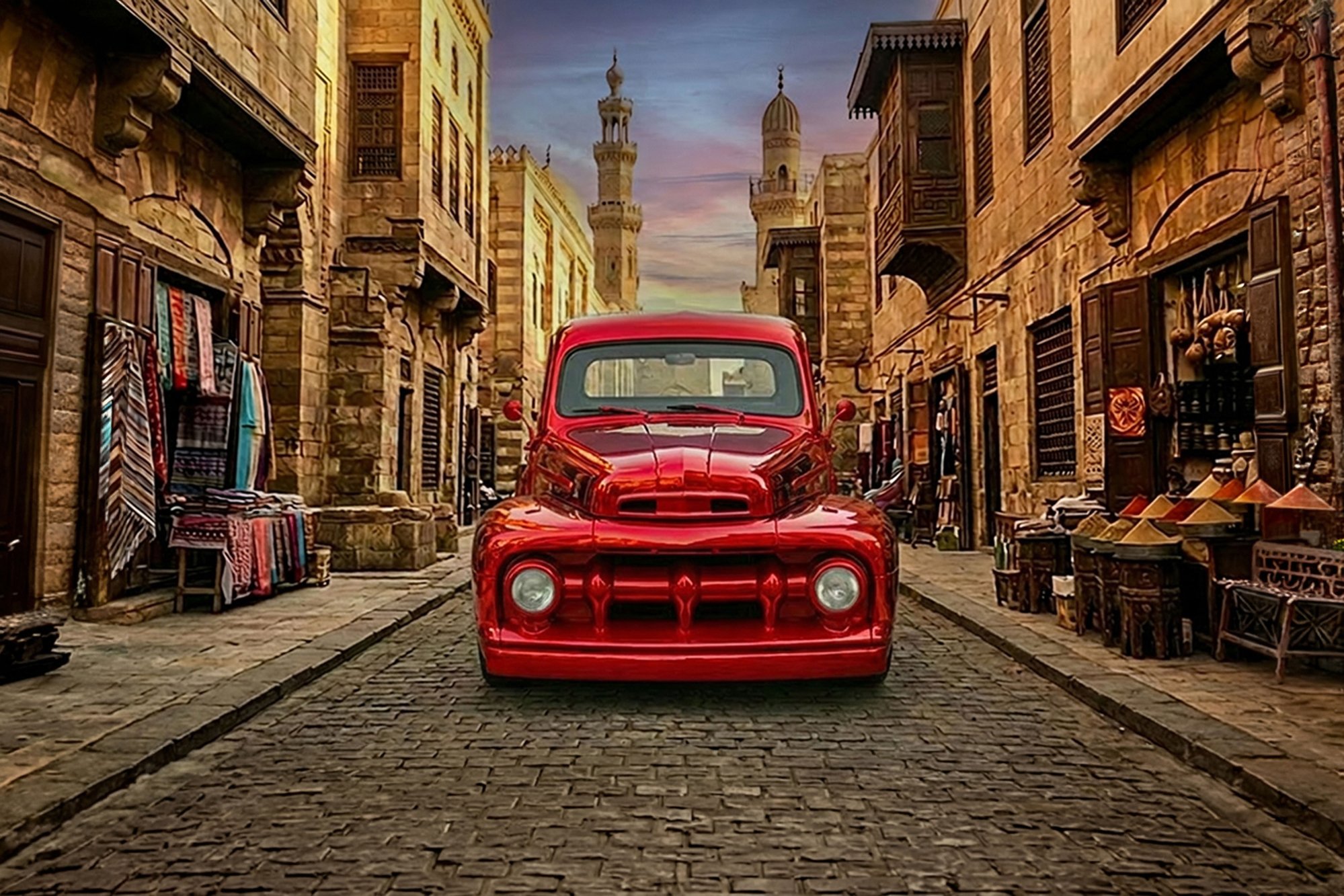 A vintage red truck parked on a cobblestone street in an old Middle Eastern town with stone buildings, narrow alleyways, and minarets in the background during sunset.