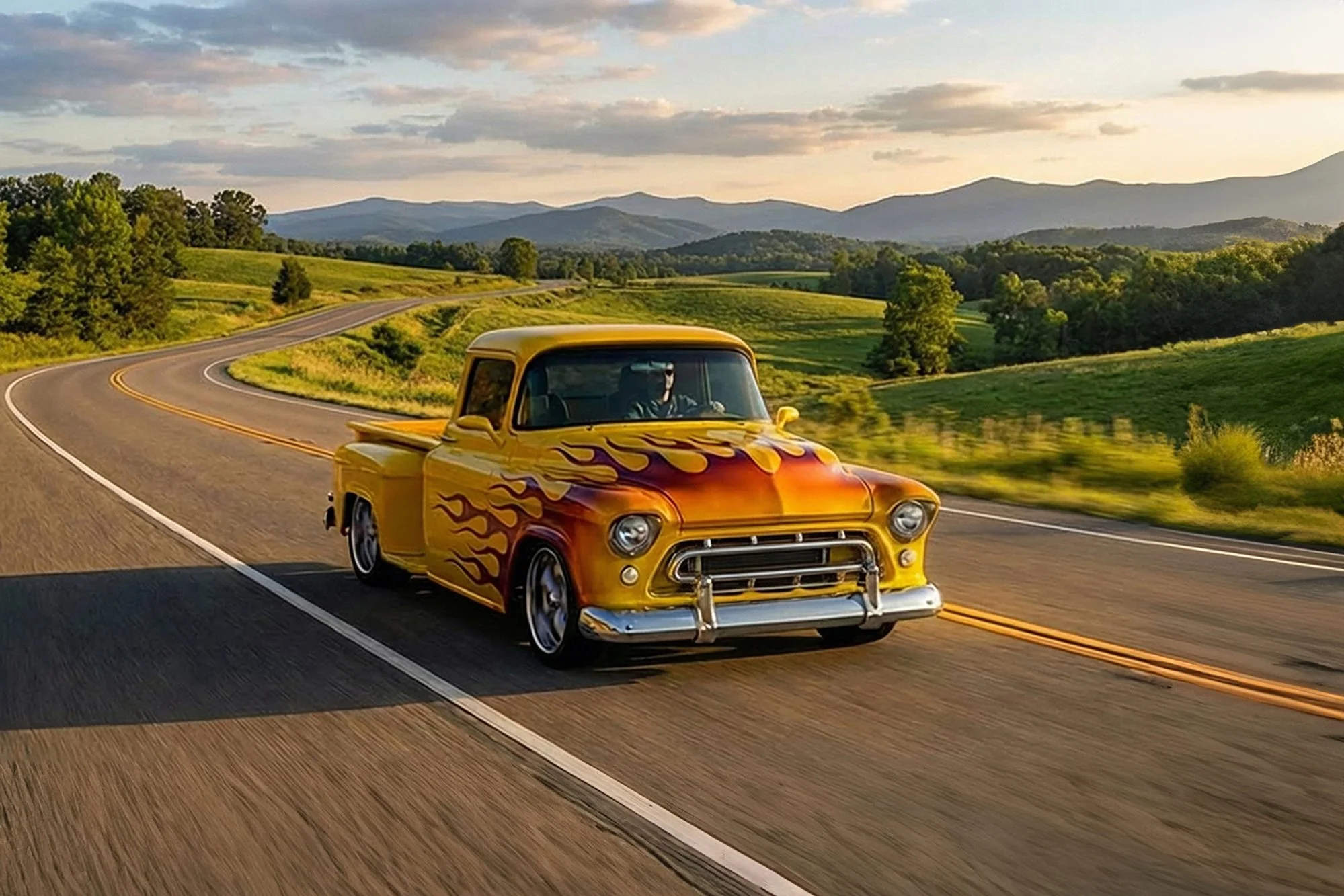 A yellow vintage pickup truck with fire flame decals driving on a winding highway with green fields and mountains in the background during sunset.