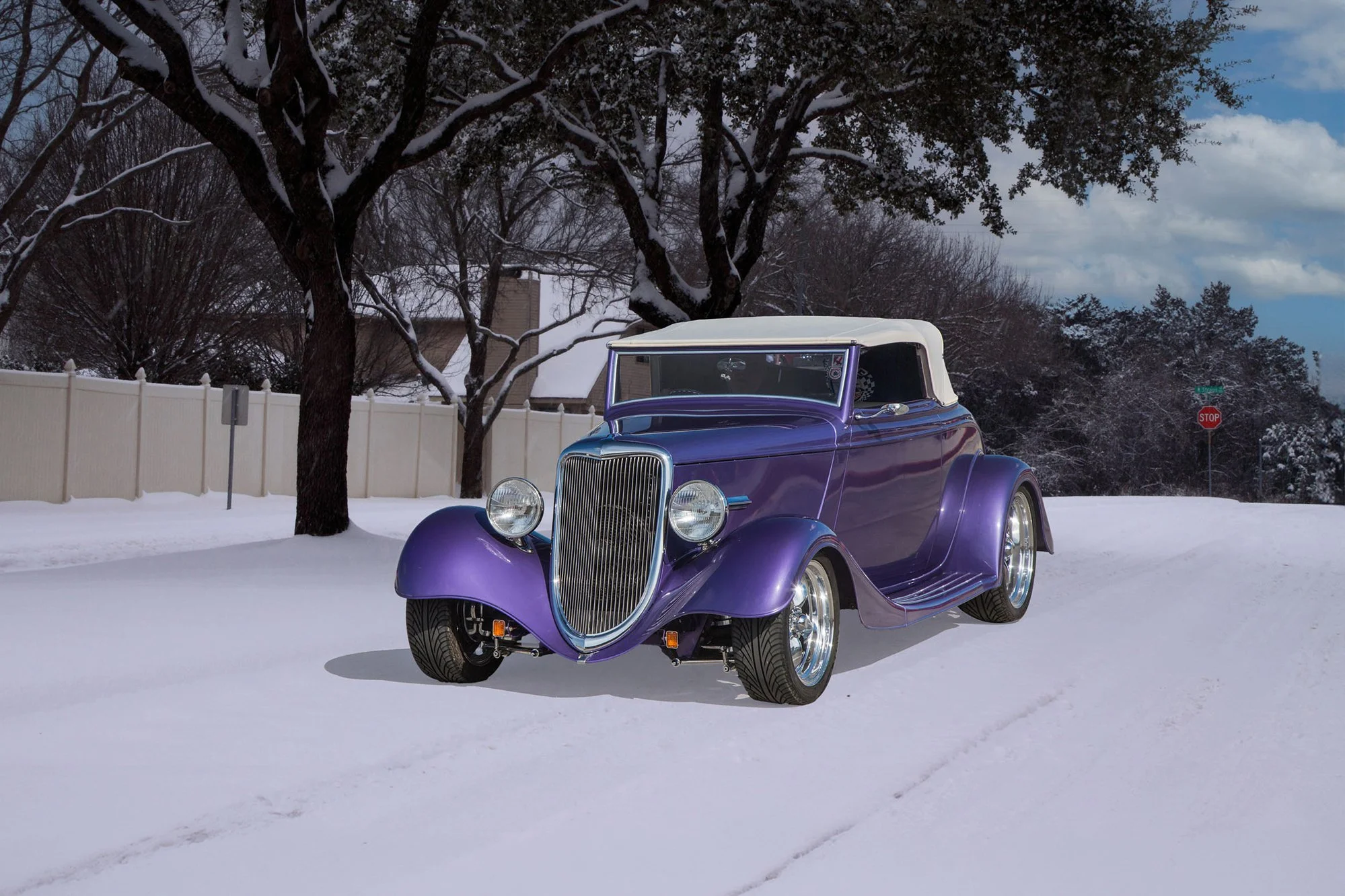 A vintage purple car with a white roof parked in a snow-covered yard with leafless trees, a white fence, and a cloudy sky in the background.