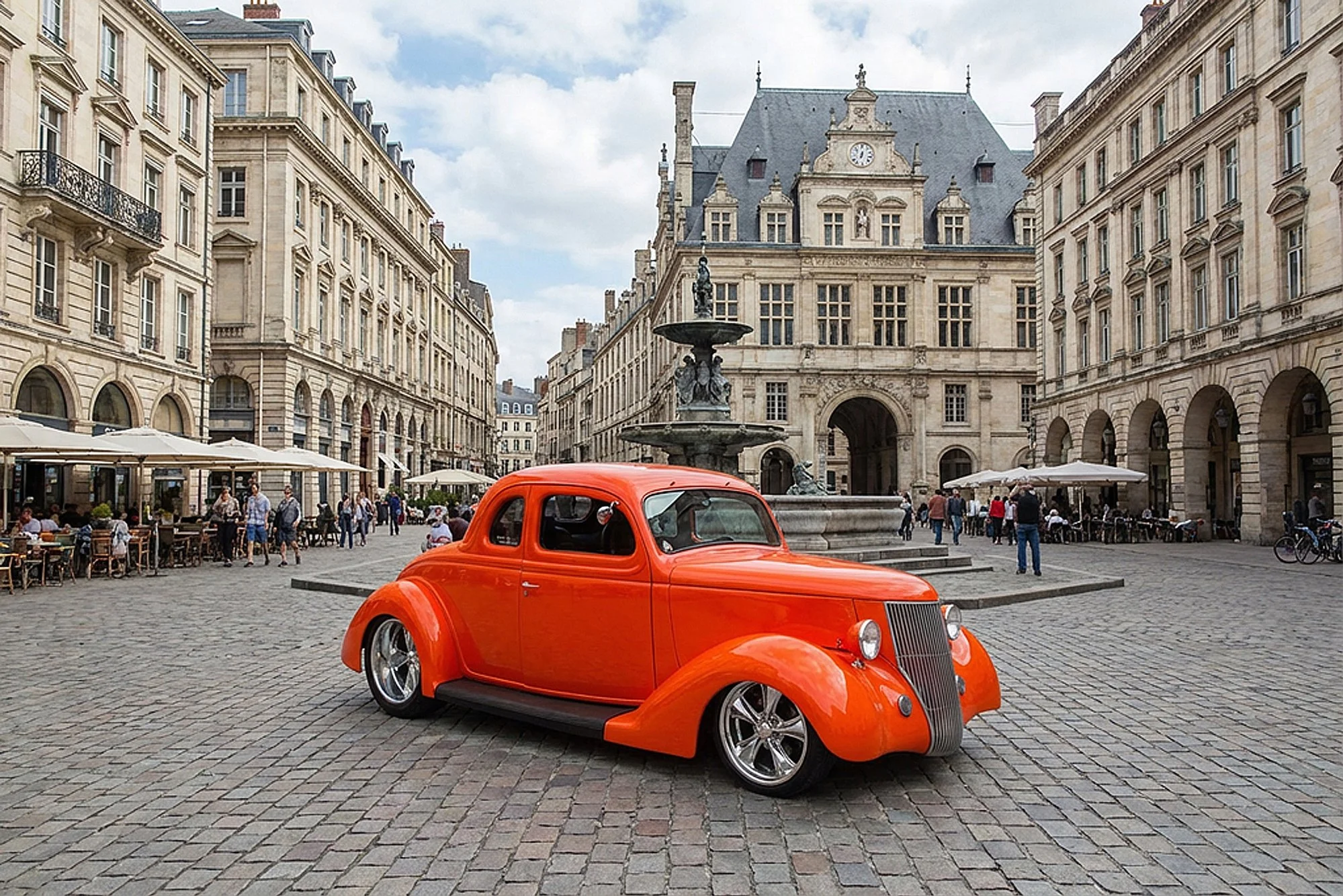 A bright orange vintage car parked in a European city square with classic architecture, a fountain, outdoor cafes with people, and a cloudy sky.