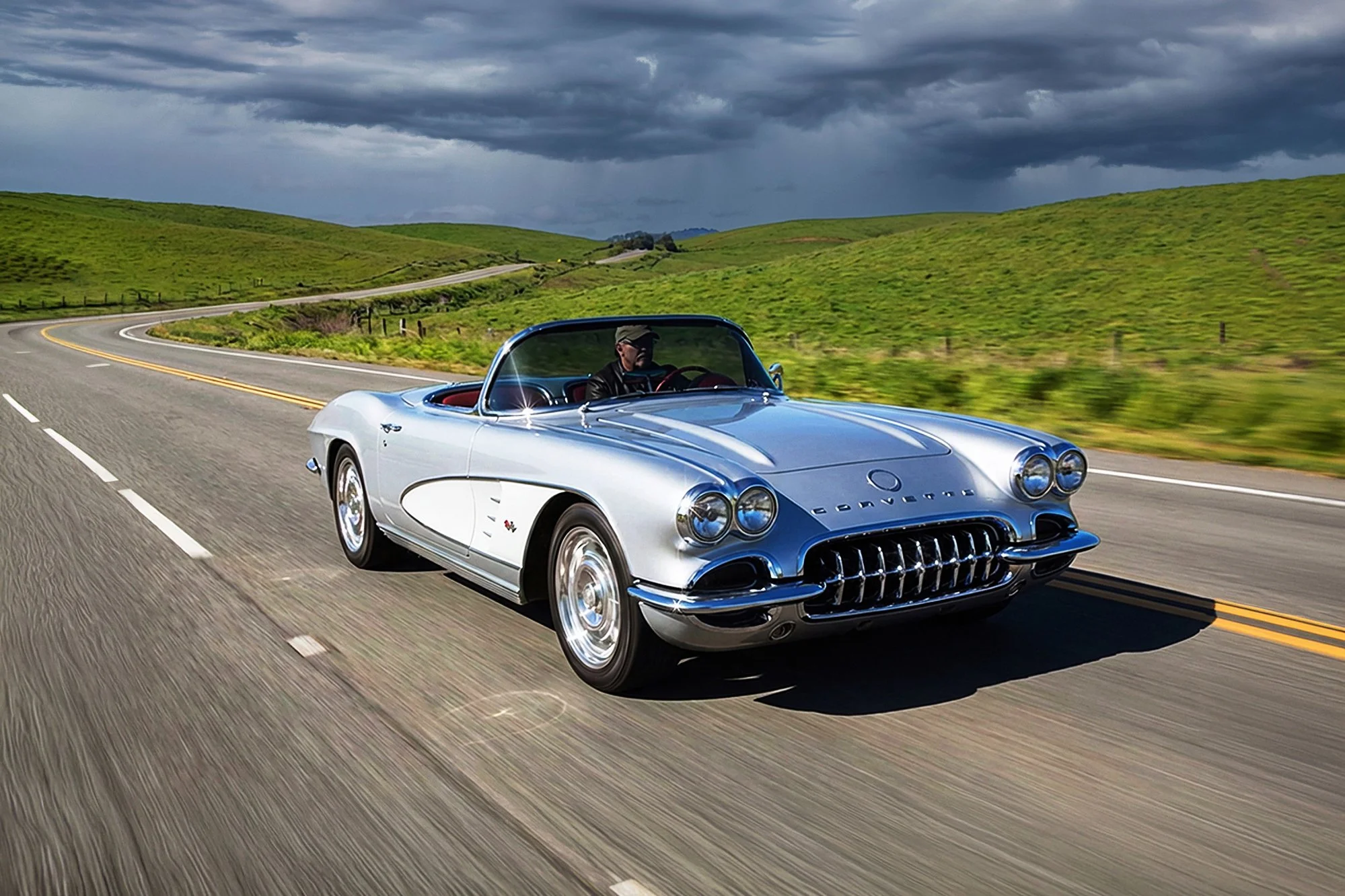 A vintage silver Chevrolet Corvette convertible driving on a winding road through green hills under dark, stormy clouds.