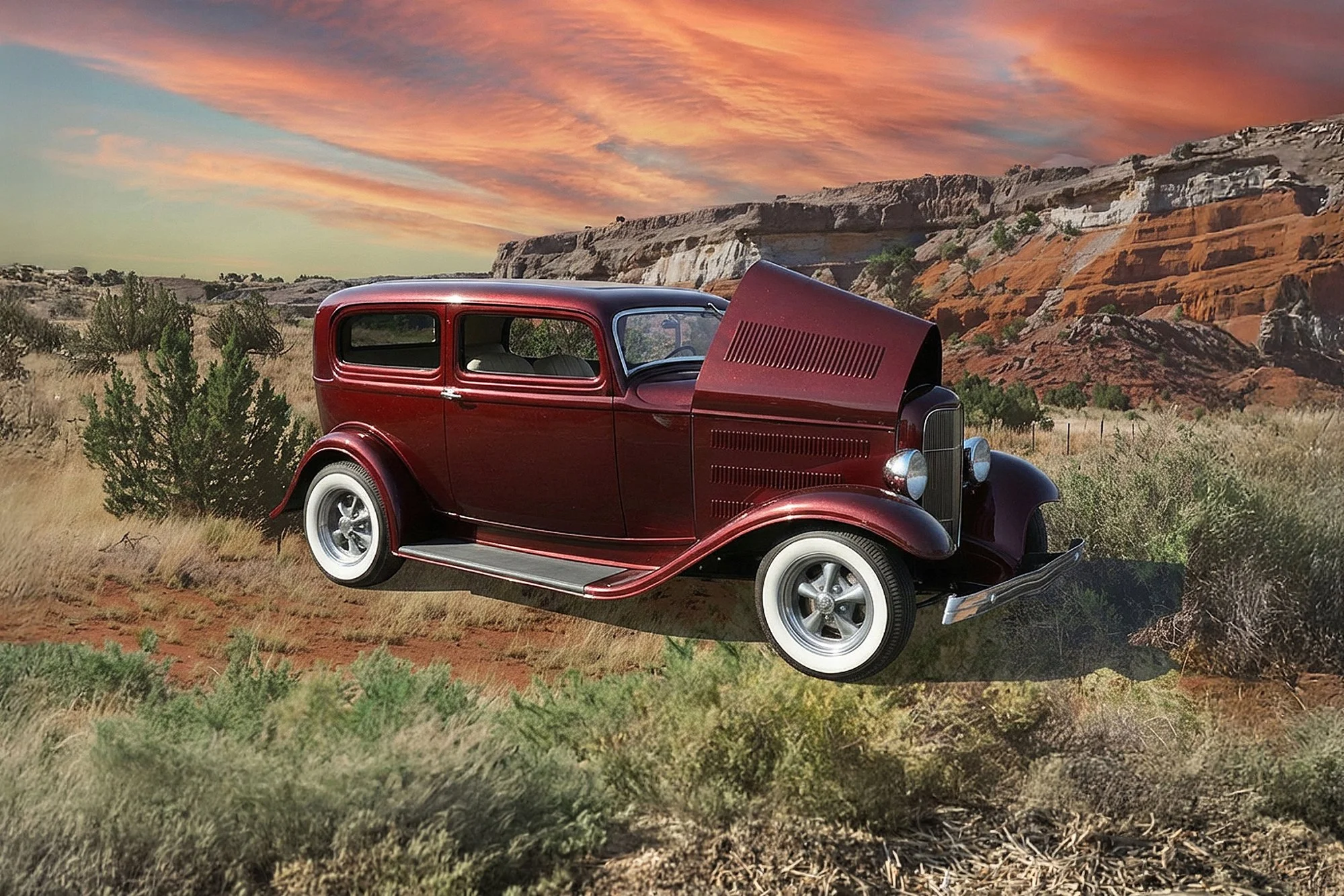 A vintage red car with its hood open, parked in a desert landscape at sunset with rocky cliffs and a colorful sky.