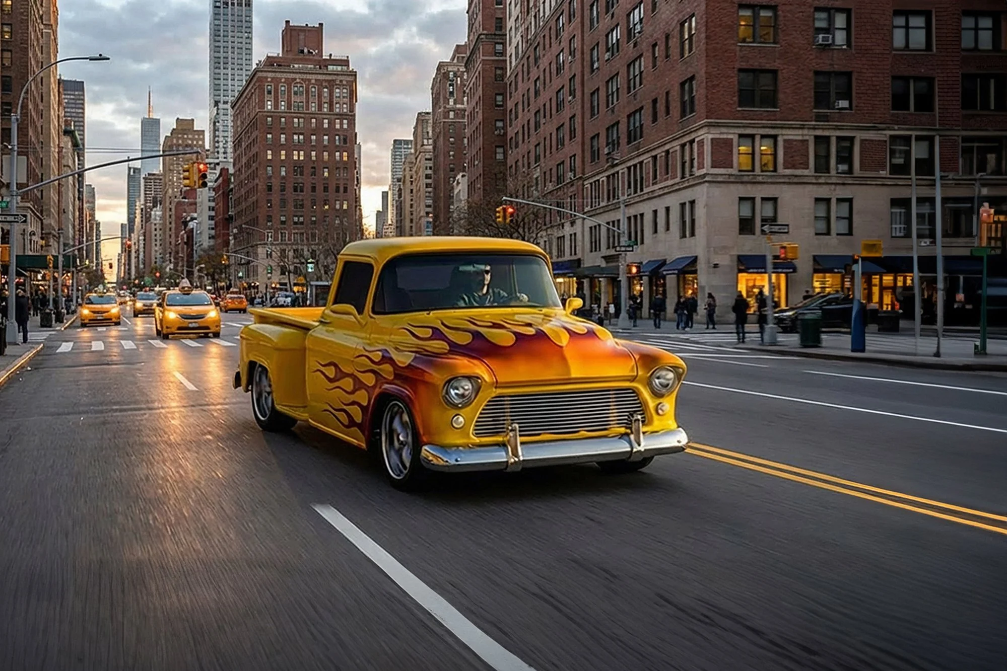 A yellow vintage pickup truck with red and orange flames painted on the front and sides drives down a city street lined with tall red brick buildings, with traffic lights and cars in the background during an overcast evening.