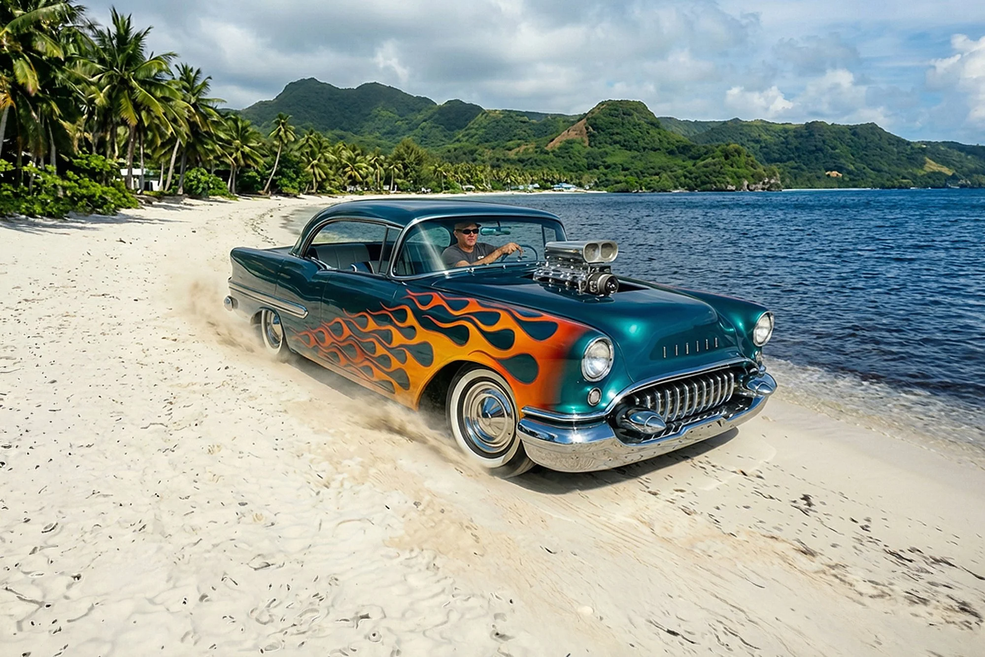 A vintage blue car with flame decals on the side driving on a sandy beach near a body of water, with lush green hills and palm trees in the background.